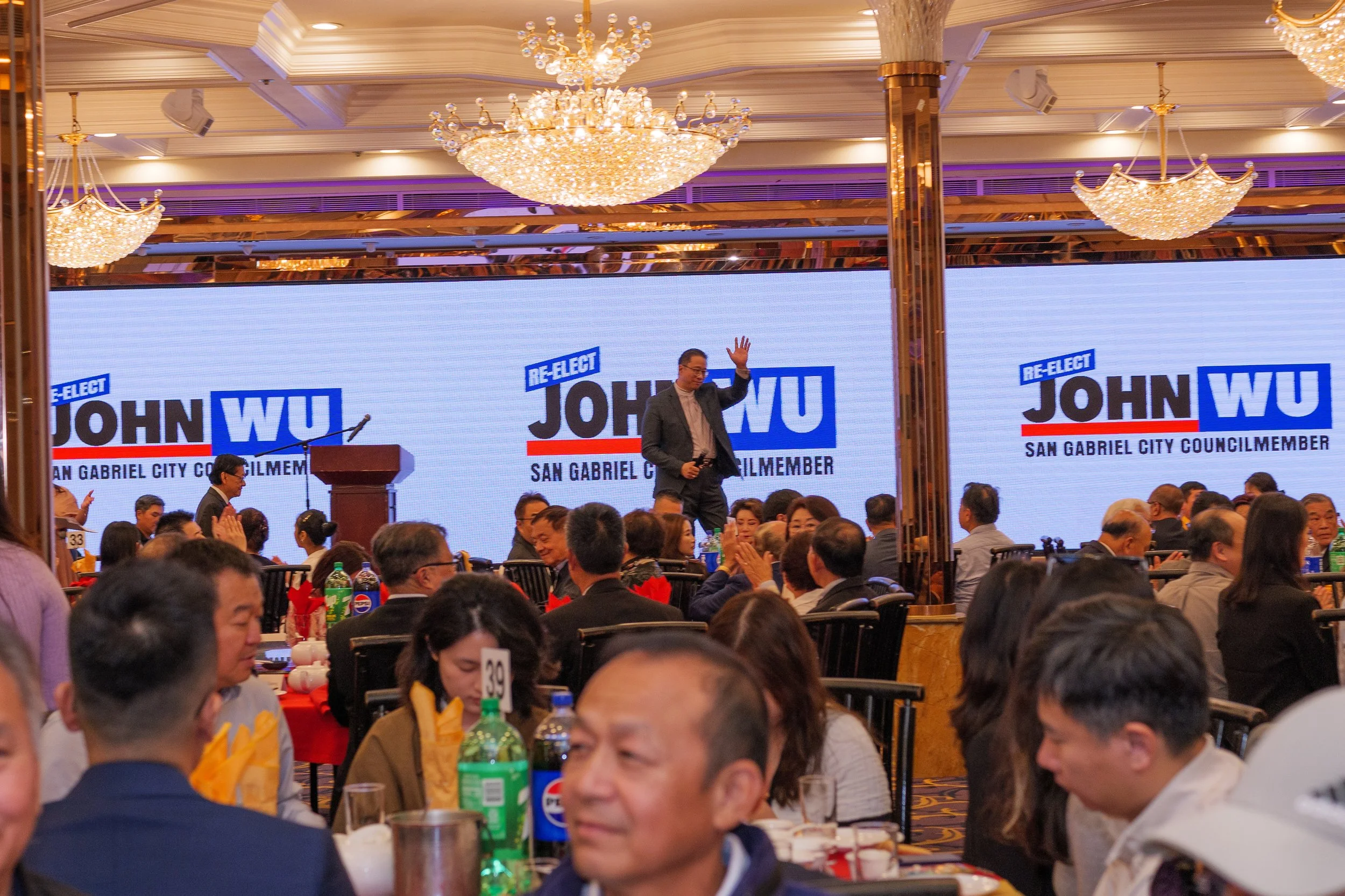 A man standing on stage at a large banquet with a background displaying campaign signs for John Wu, a San Gabriel City Councilmember, waving to an audience of mostly seated people enjoying a formal event with chandeliers and tables.