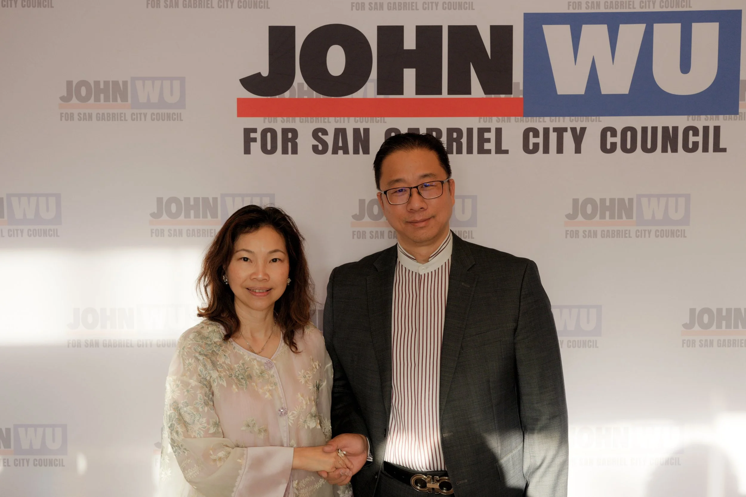 Two people, a woman and a man, standing together and shaking hands in front of a backdrop that says 'John Wu for San Gabriel City Council.' The woman has wavy hair and is wearing a light-colored, floral-embroidered traditional outfit, while the man h