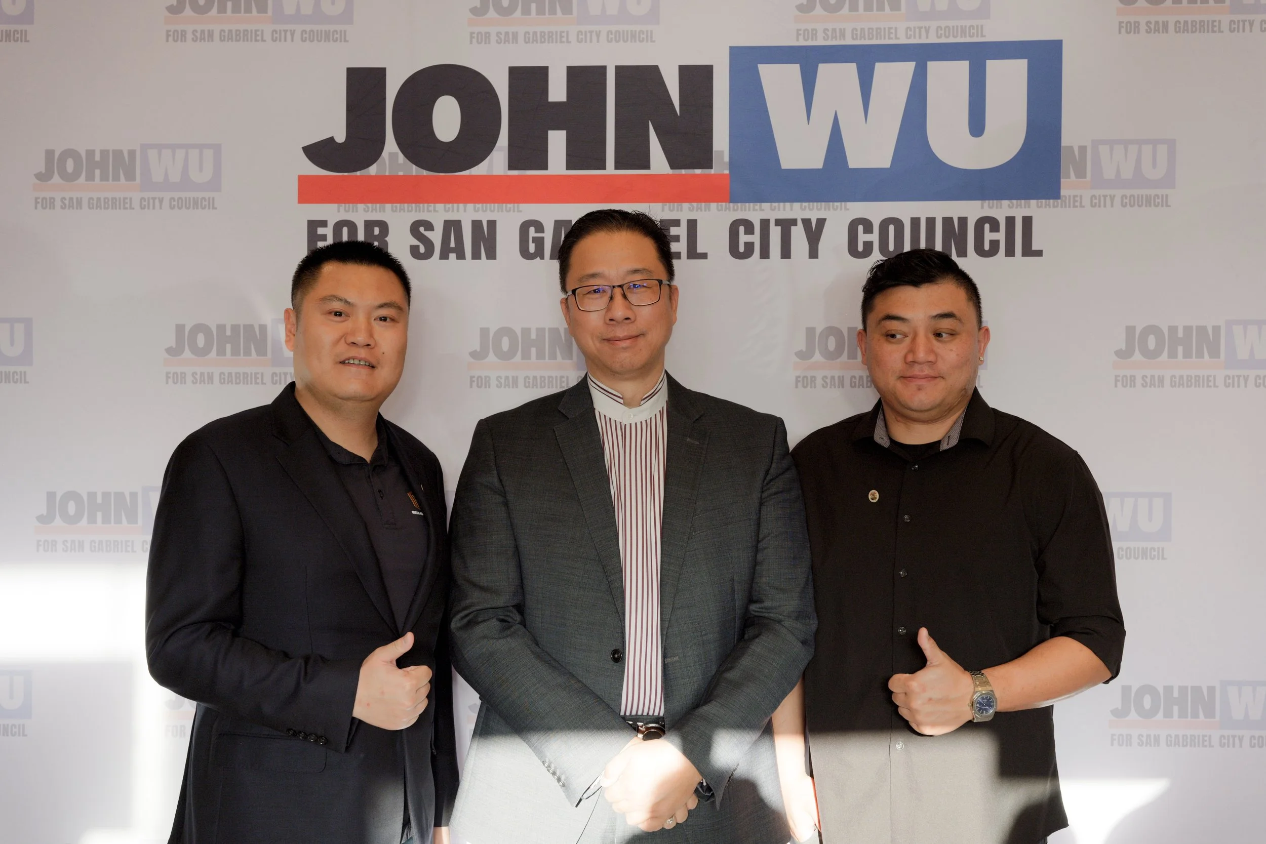 Three men in formal attire standing together, smiling, in front of a backdrop with campaign logos for John Wu for San Gabriel City Council.