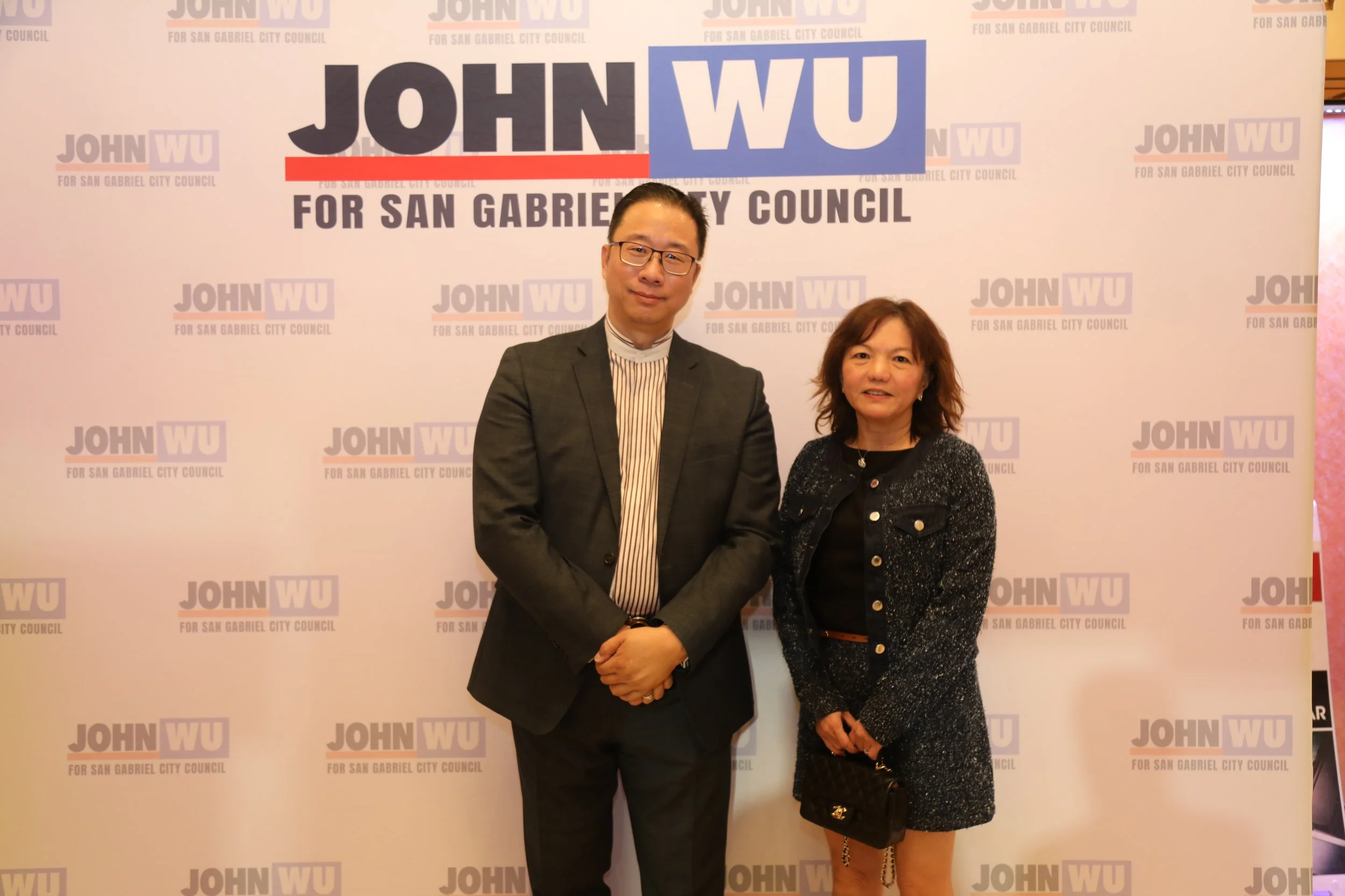 A man and a woman are standing in front of a backdrop that says 'John Wu for San Gabriel City Council' repeatedly. They are shaking hands, and both are smiling at the camera. The man is wearing glasses and a suit, and the woman is wearing a black jac