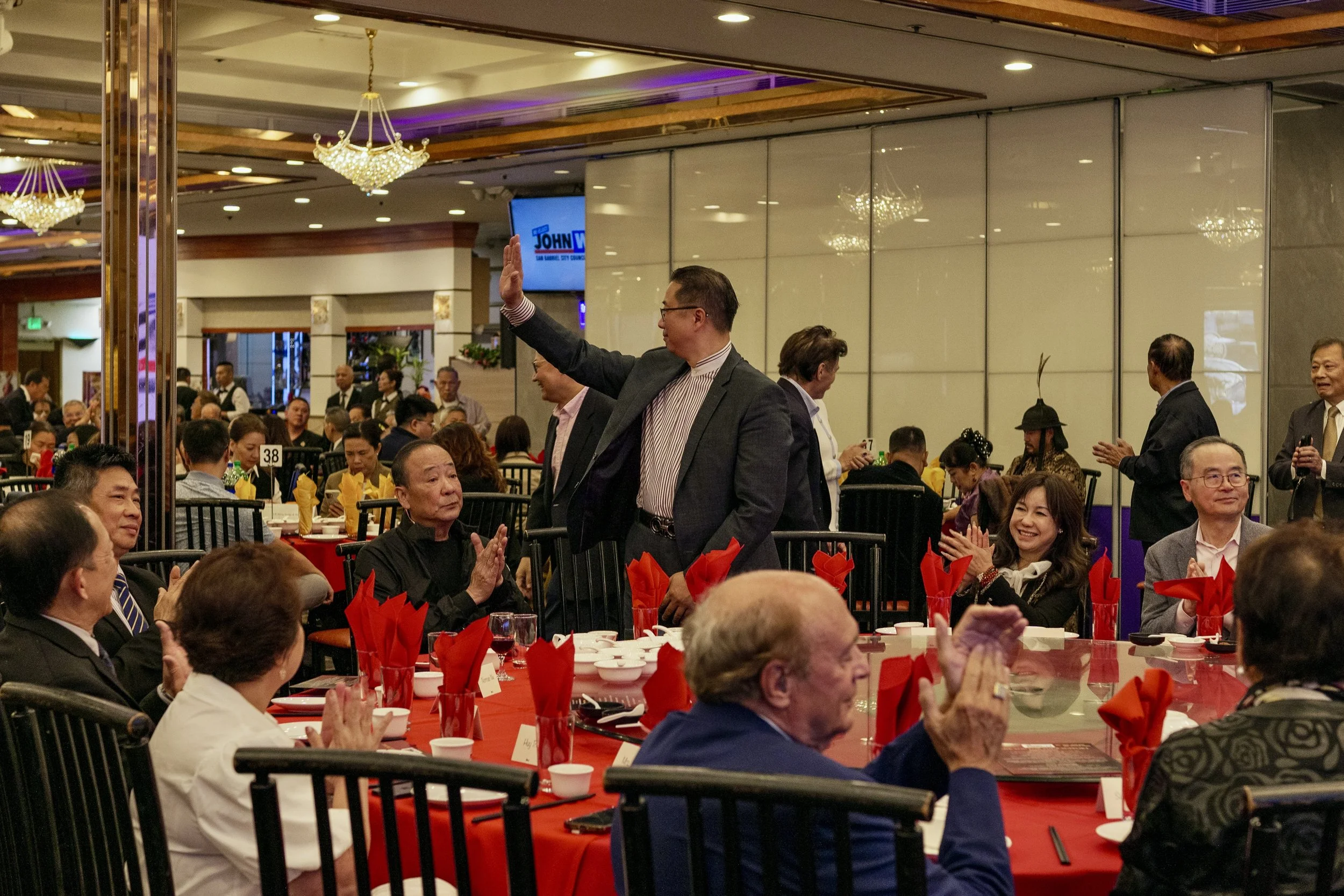 A man in a suit with glasses is standing and waving at a large banquet or dinner event in a decorated hall. The hall has chandeliers, a mirror wall, and tables with people seated, clapping, and smiling. The tables are set with red napkins, white cups