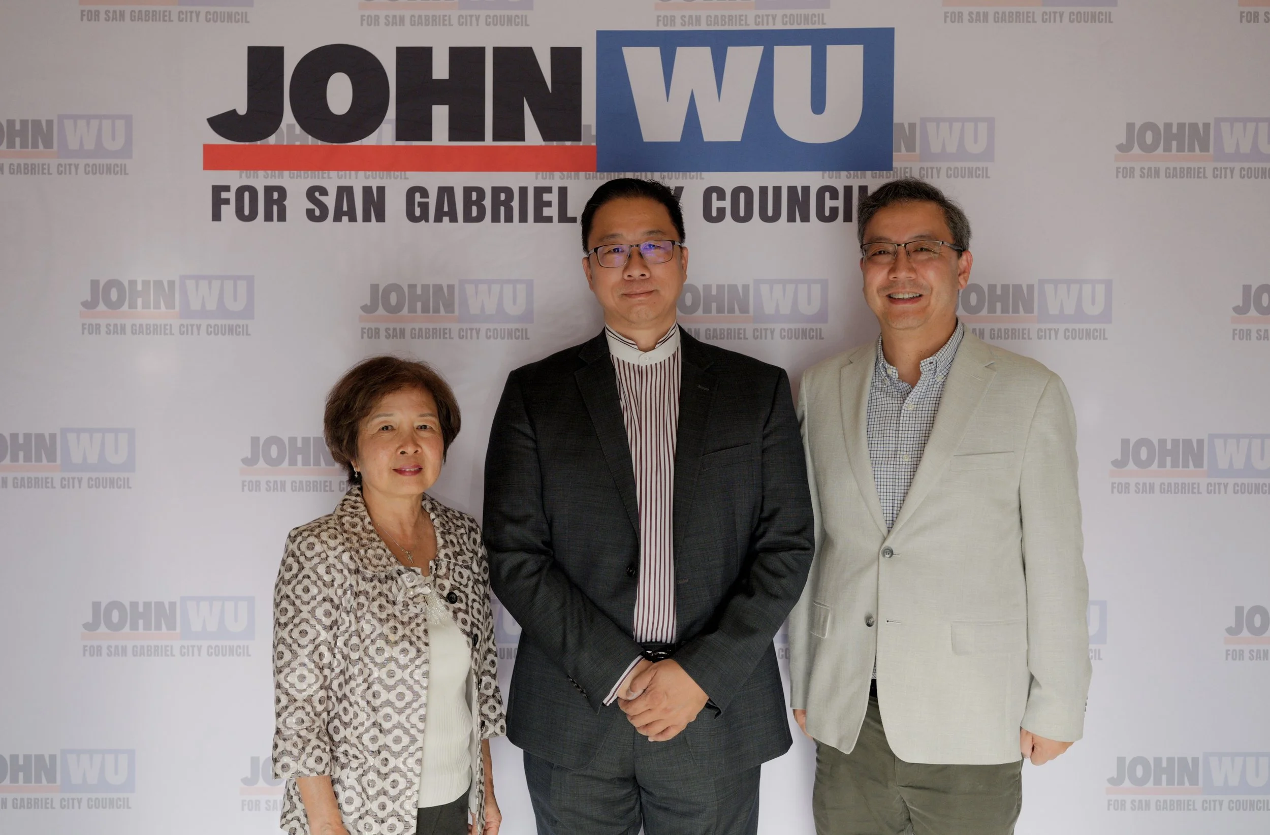 Three people standing in front of a backdrop with the words 'John Wu for San Gabriel City Council.' The woman on the left is wearing a leopard print blazer, and the two men are in suits, with one wearing glasses and the other smiling.
