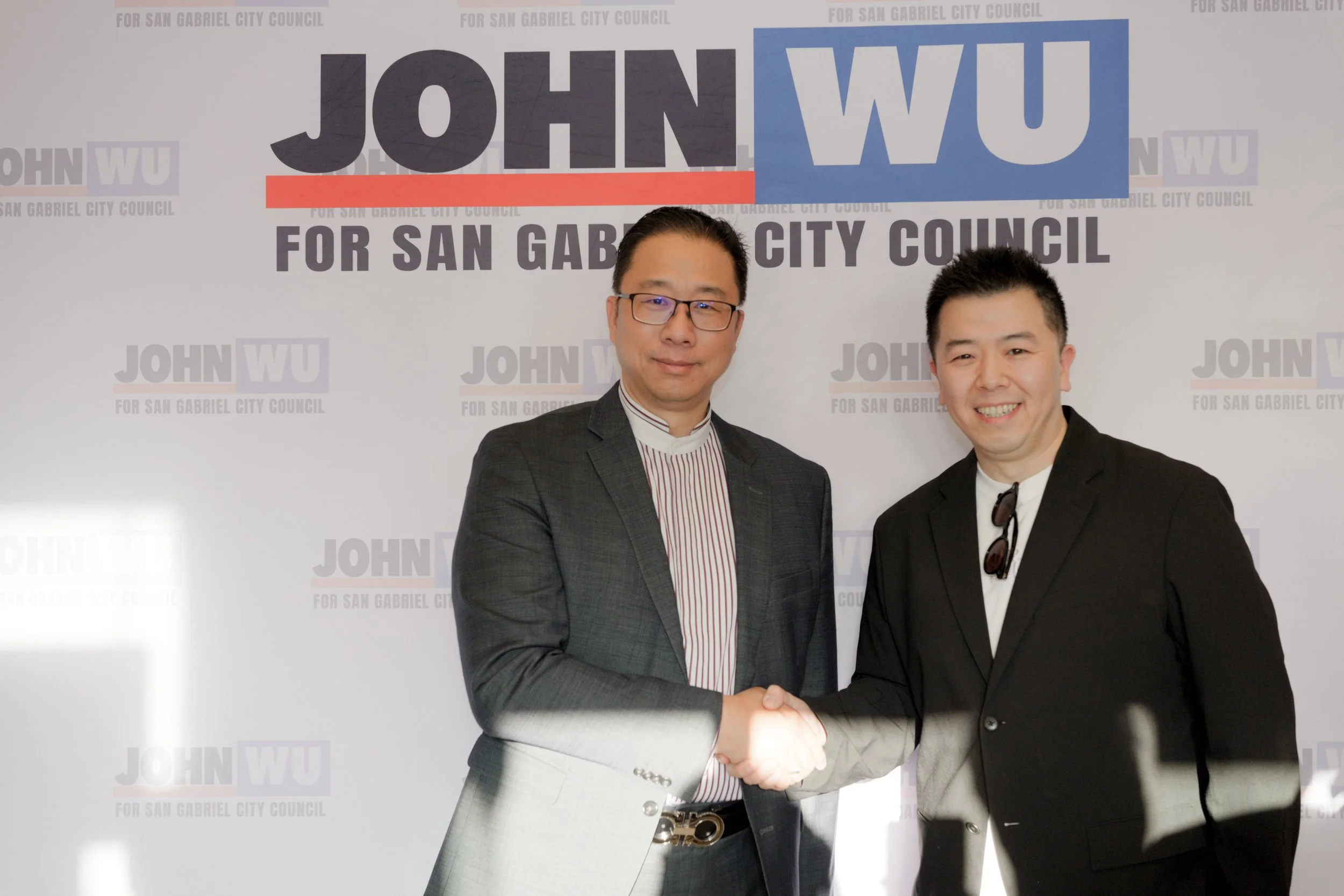 Two men in suits shaking hands at a political campaign event, standing in front of a backing with the text 'JOHN WU for San Gabriel City Council'.