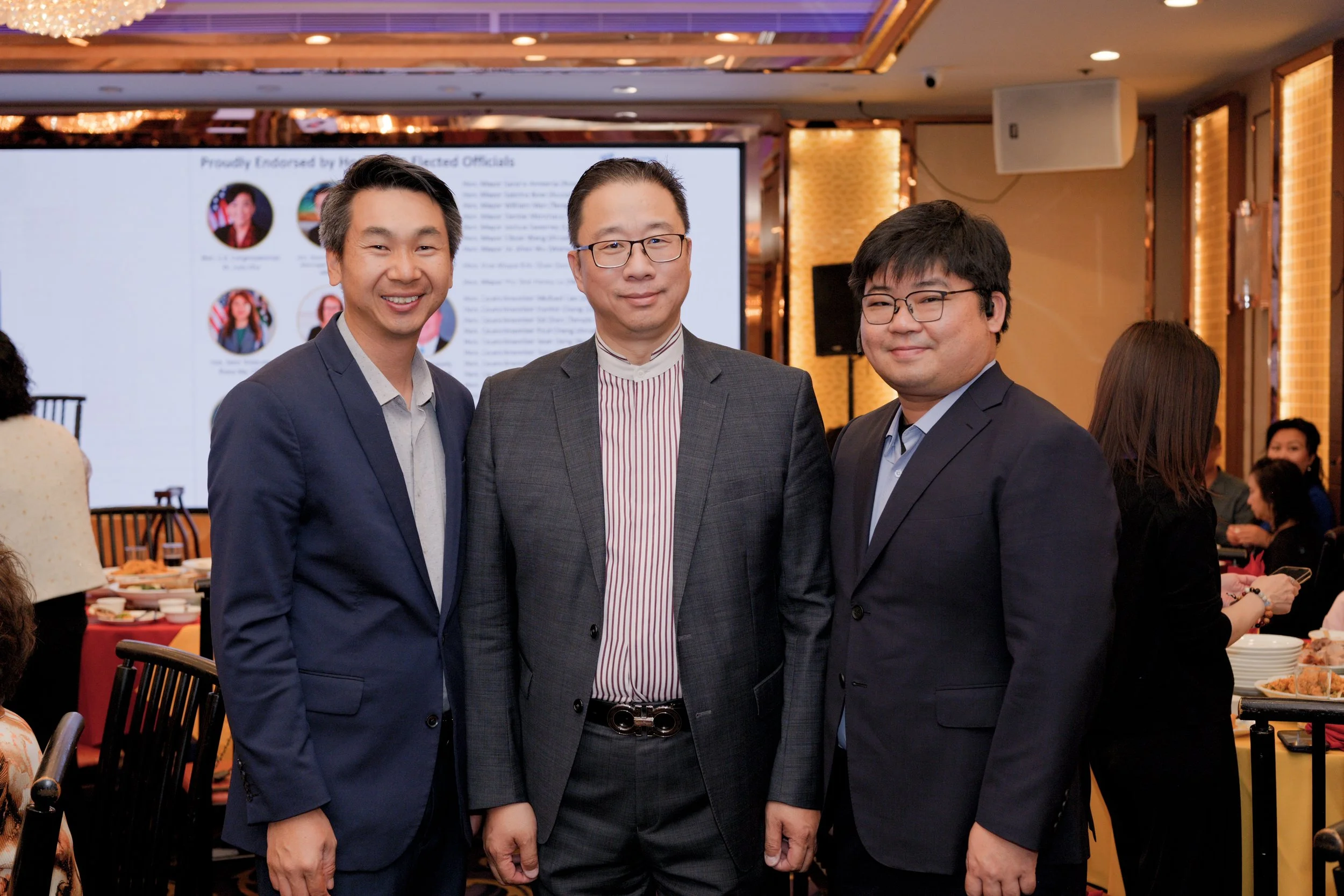 Three men in suits posing together at a formal event, with a large screen and other guests in the background.
