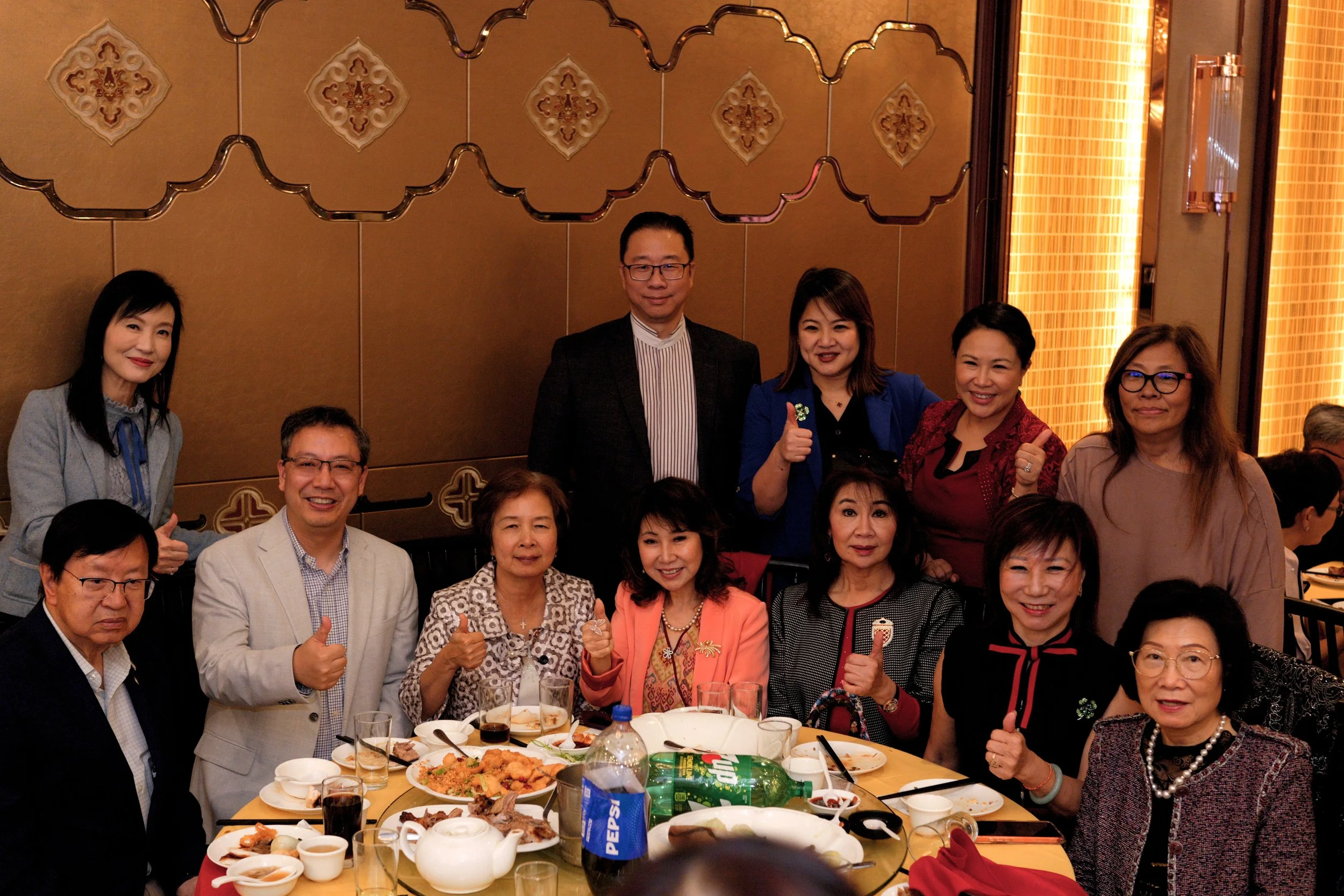A group of people gathered at a dining table in a restaurant, smiling and posing for a photo. The table is filled with various dishes, drinks, and utensils. The background features a warm-colored wall with decorative patterns and lighting.