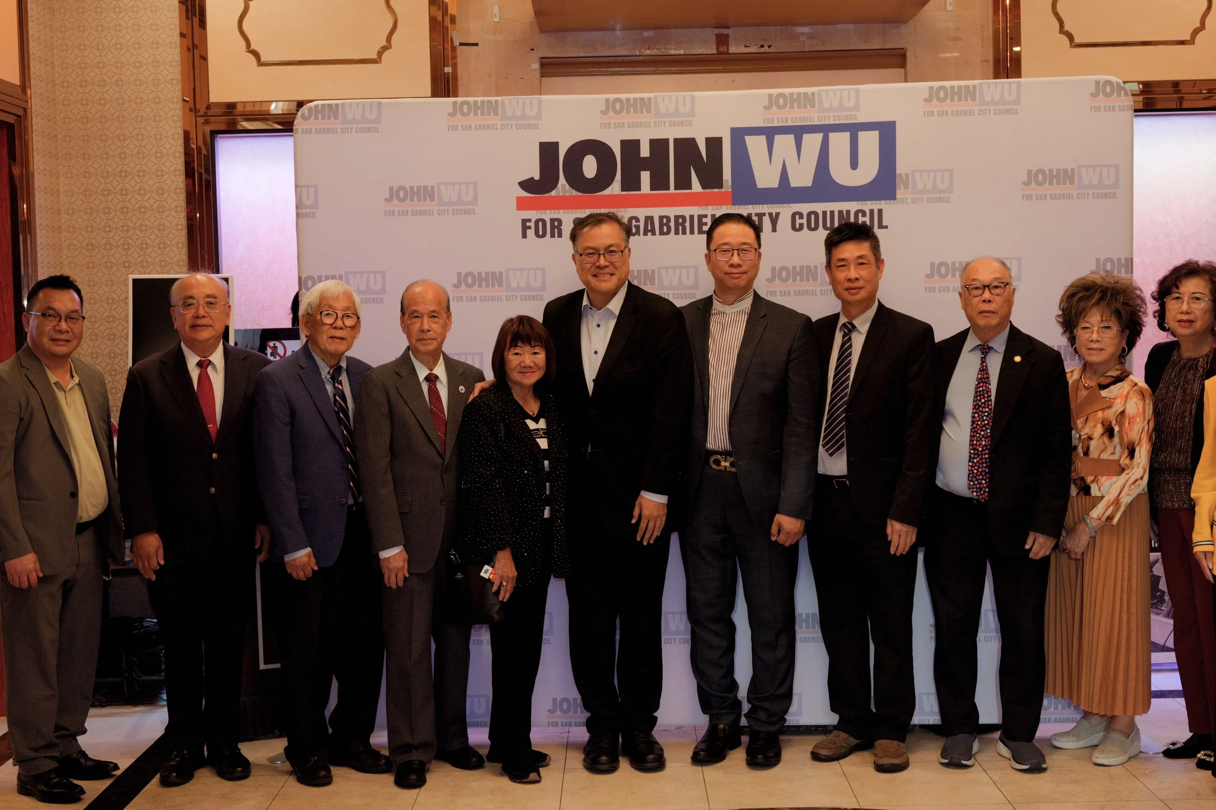 A group of eleven people, nine men and two women, standing side by side in front of a large banner that reads 'JOHN WU for San Gabriel City Council.' The people are dressed in business attire and are smiling at the camera. The background appears to b