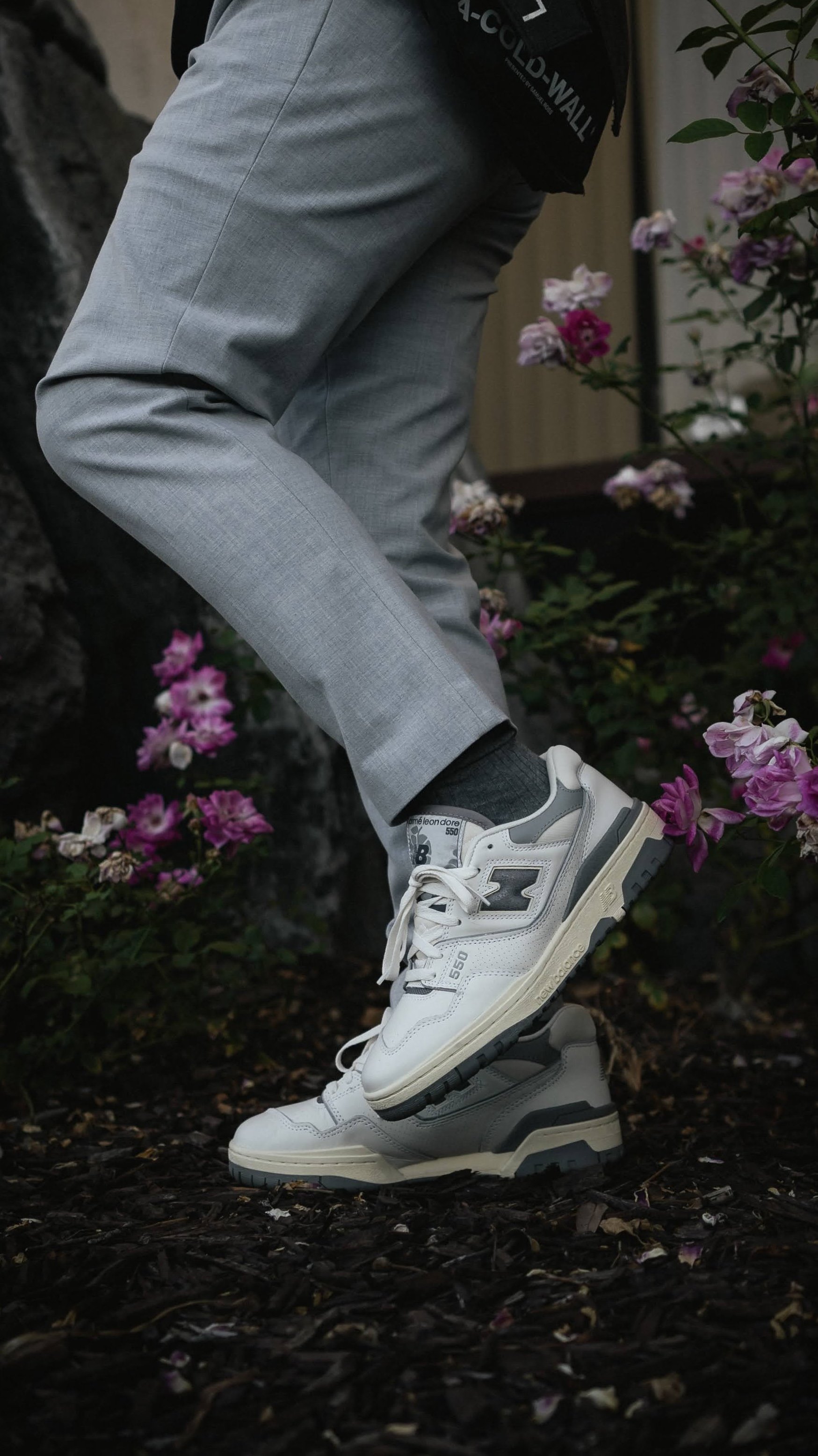 Person wearing gray plaid pants and white sneakers balancing on one foot in a garden with pink flowers.
