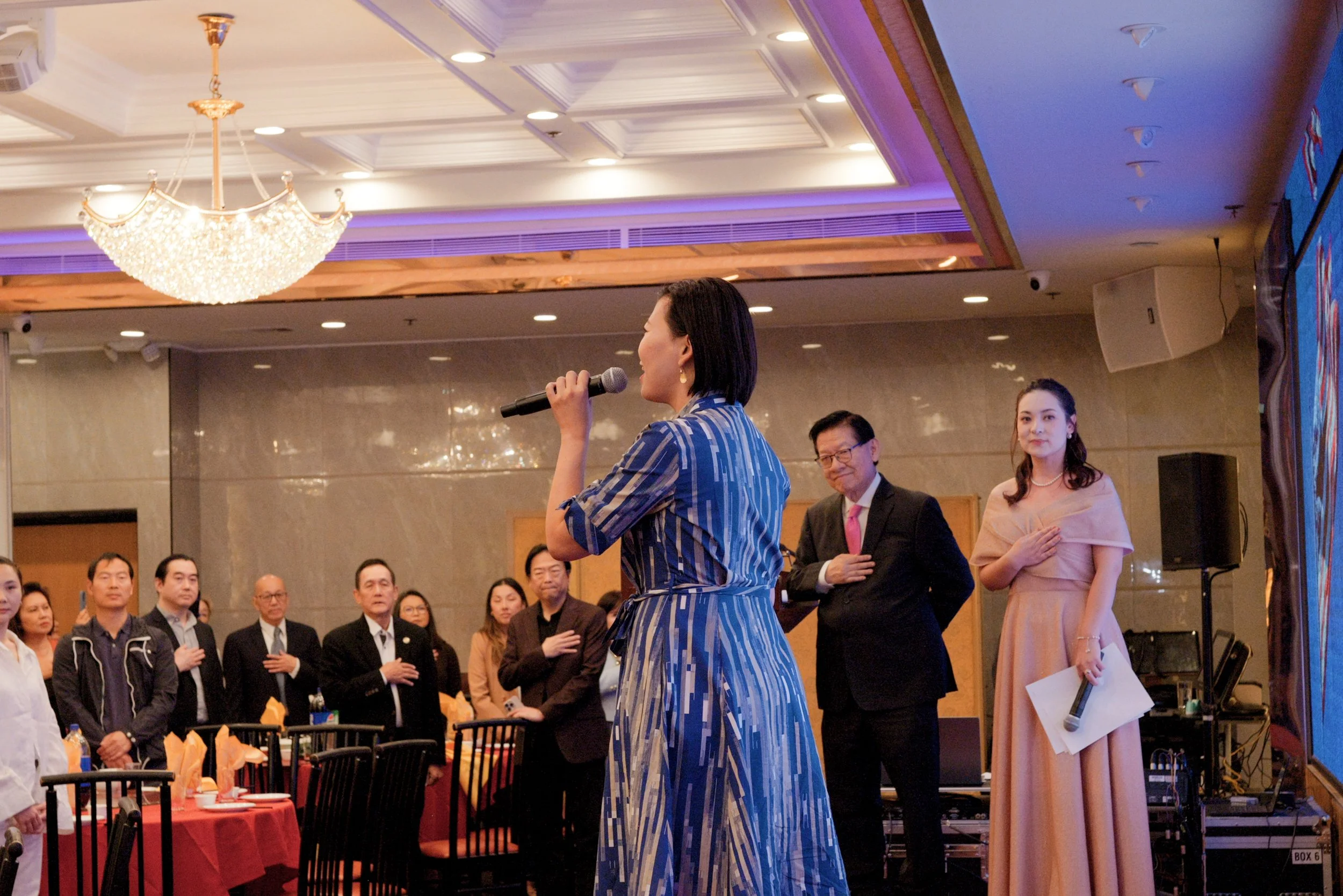 A woman in a blue and white dress holds a microphone and appears to be speaking to a crowd in an indoor event with a chandelier on the ceiling and a large screen to the right. Several people are standing attentively in the background, including a wom