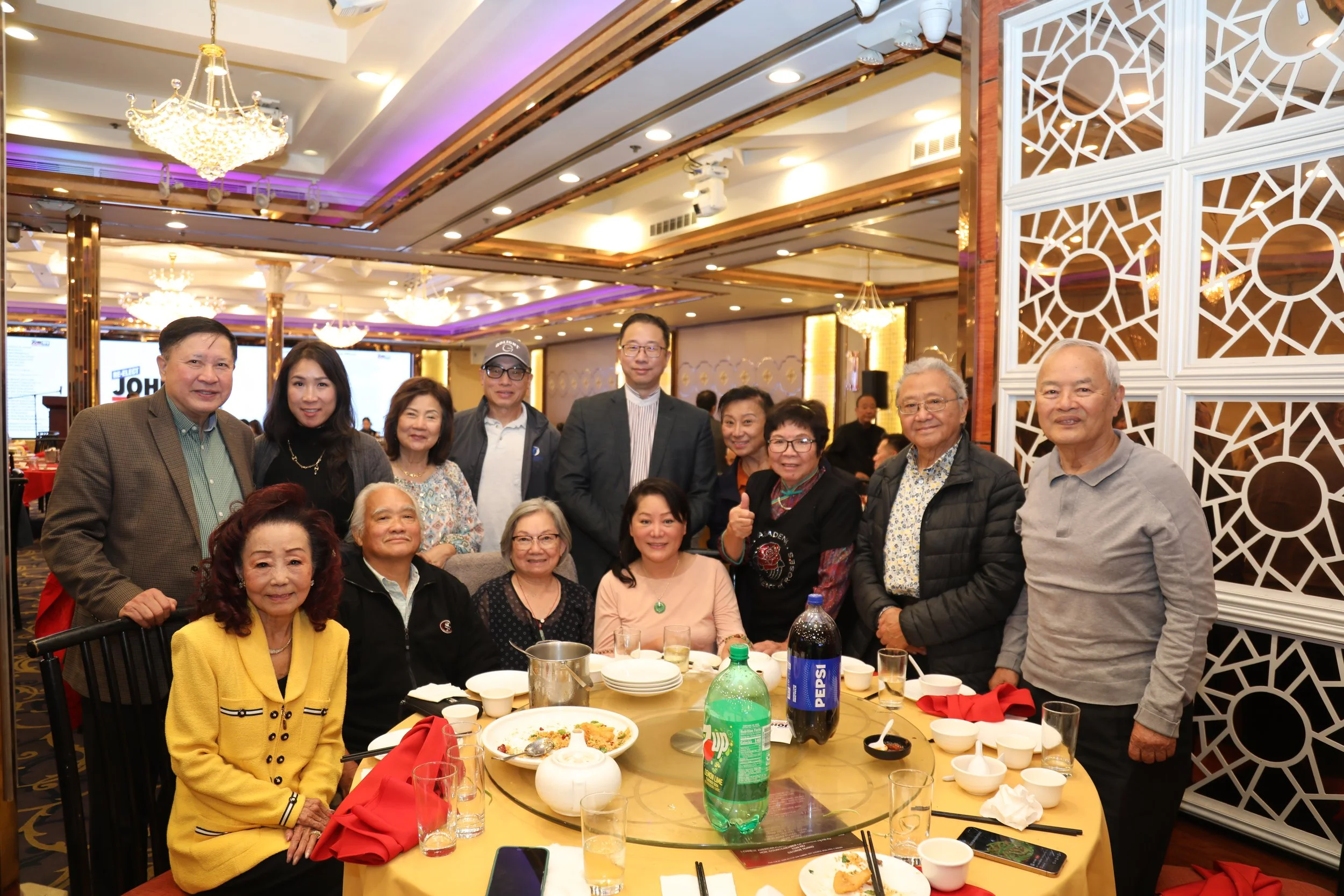 A group of elderly and middle-aged people gathered at a restaurant, standing behind and seated around a dining table with food and drinks, in a decorated banquet hall with chandeliers and ornate white wall designs.