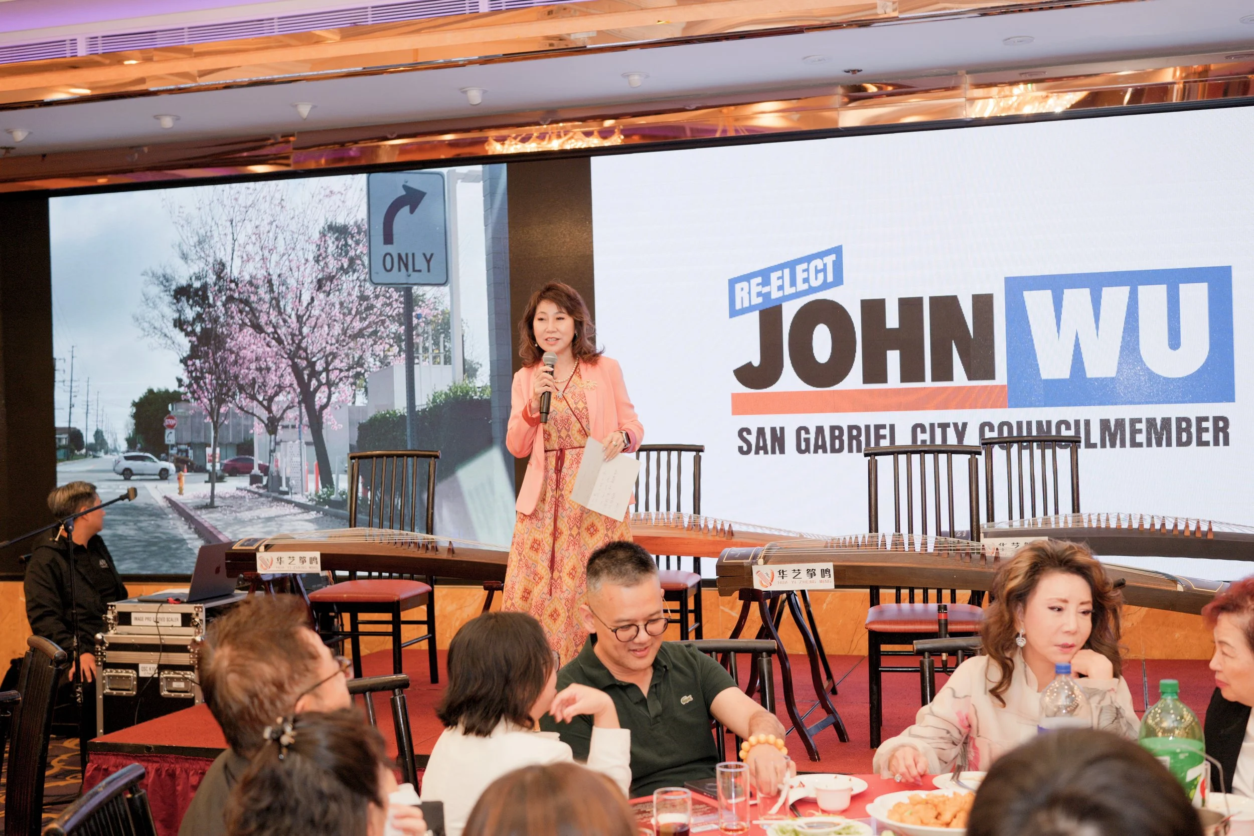 A woman in a peach-colored blazer and patterned dress is speaking into a microphone on a stage. Behind her, a large screen displays a campaign sign for John Wu, a San Gabriel City Council member, with a background of cherry blossom trees.