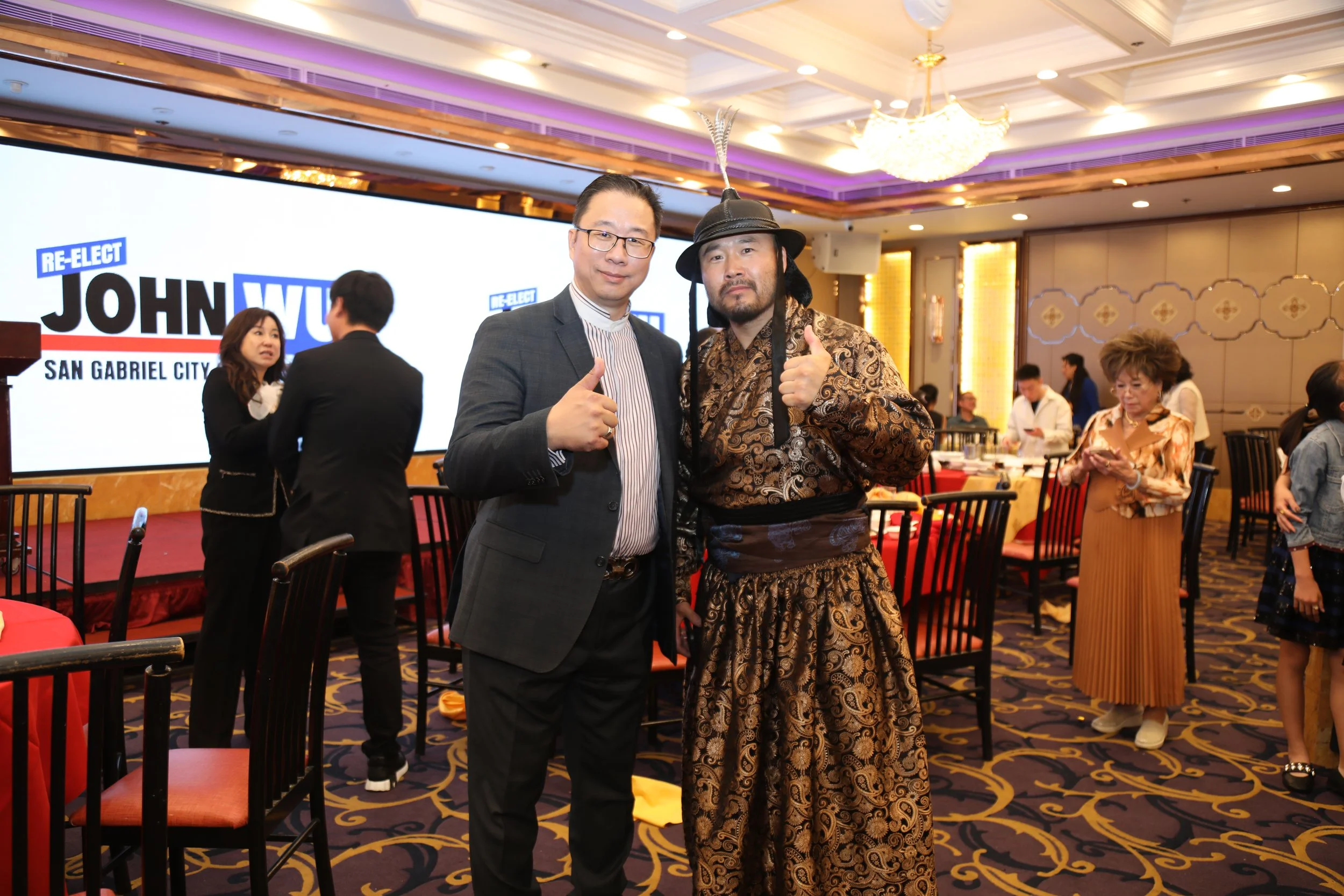 Two men in the foreground at a conference, one in a suit giving a thumbs up and the other in a traditional costume with a feathered hat. Background shows a large screen with 'Re-Elect John Wu for San Gabriel City' and other people mingling at tables.