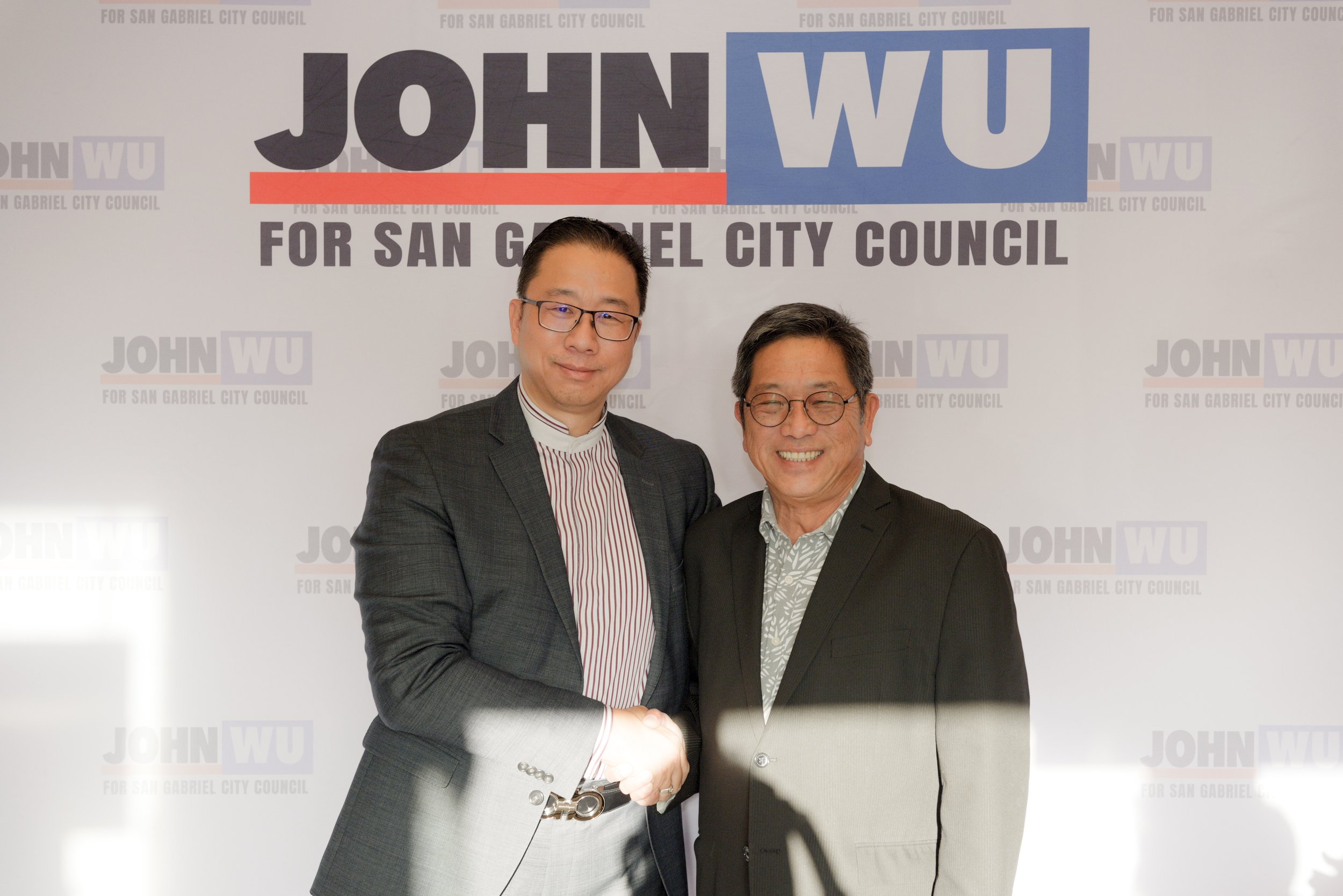 Two men shaking hands in front of a campaign backdrop that reads "JOHN WU" and "FOR SAN GABRIEL CITY COUNCIL." The man on the left is wearing glasses, a dark suit, and a striped shirt. The man on the right is smiling, wearing glasses, and a dark blaz