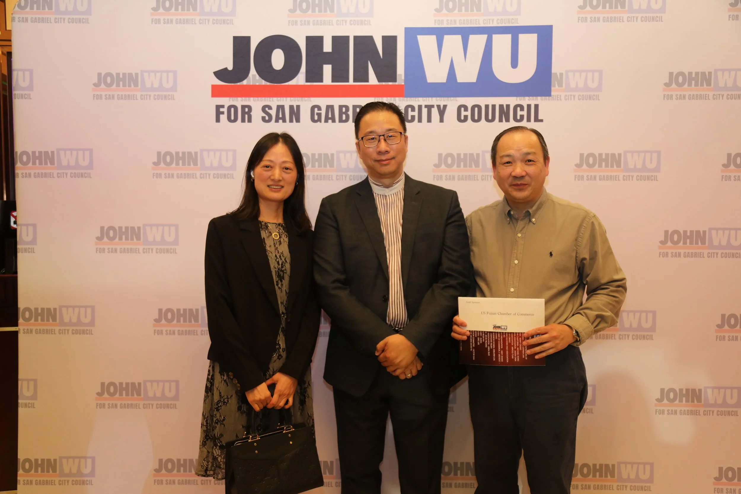 Three individuals standing in front of a backdrop with the text "JOHN WU FOR SAN GABRIEL CITY COUNCIL" repeated throughout. They are posing for a photo, with the man on the right holding a certificate.