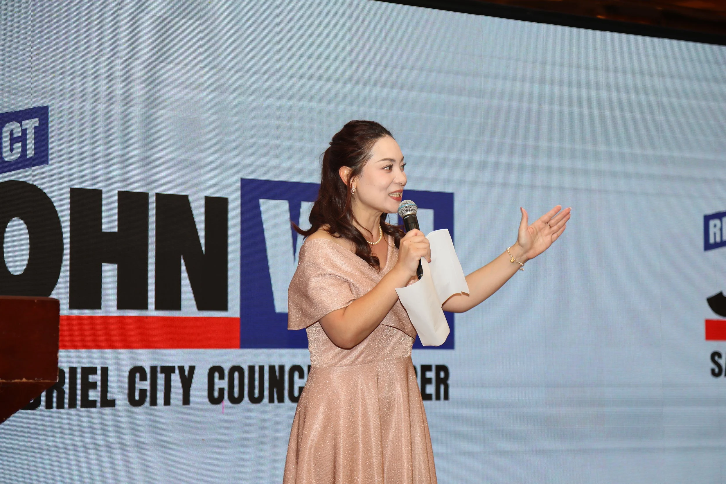 A woman in a peach-colored dress speaking at a microphone on stage during a city council event, with a large screen behind her displaying text and graphics.