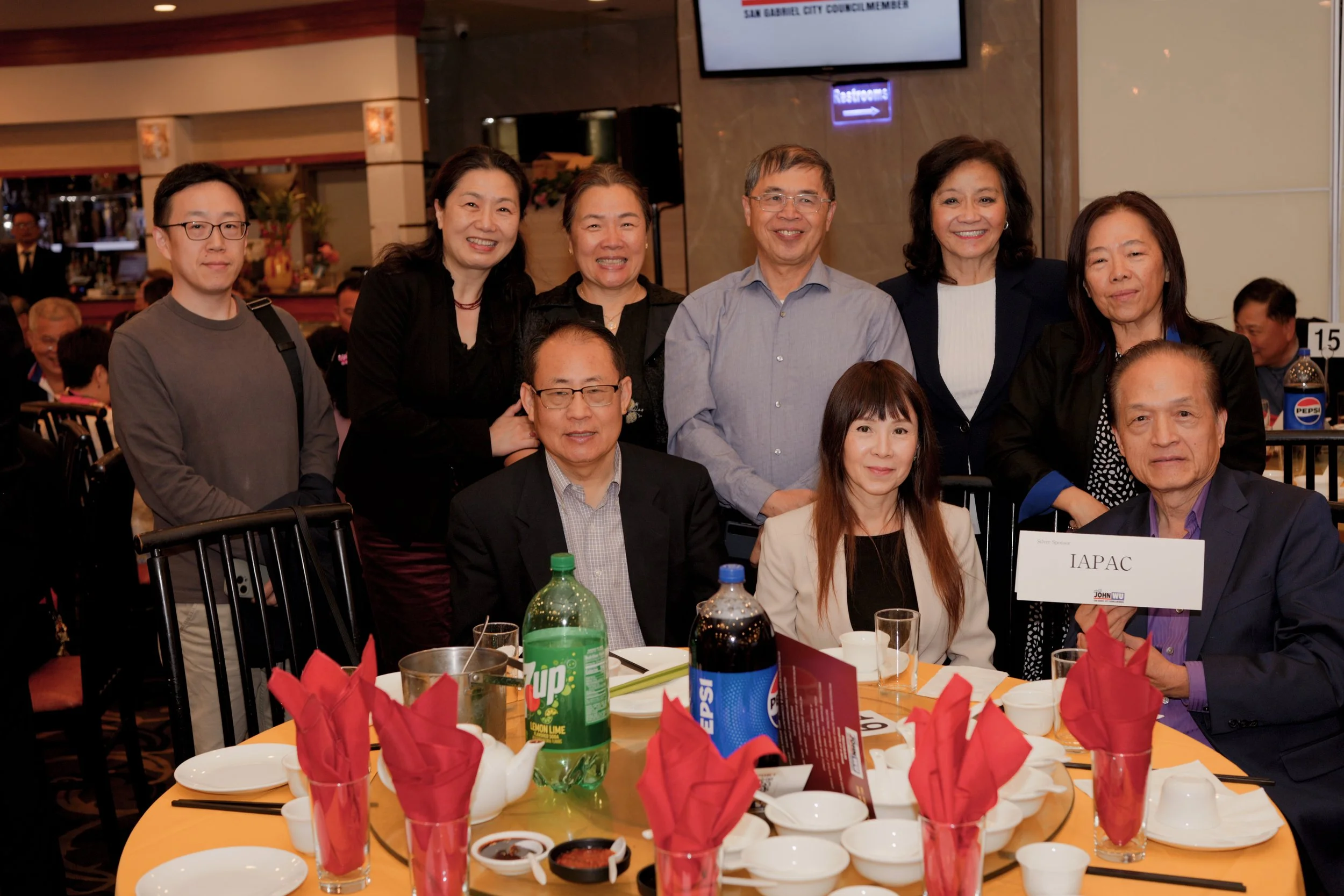 Group of eight diverse people, five women and three men, smiling and posing behind a dining table at a formal event in a restaurant or banquet hall, with food and beverages on the table.