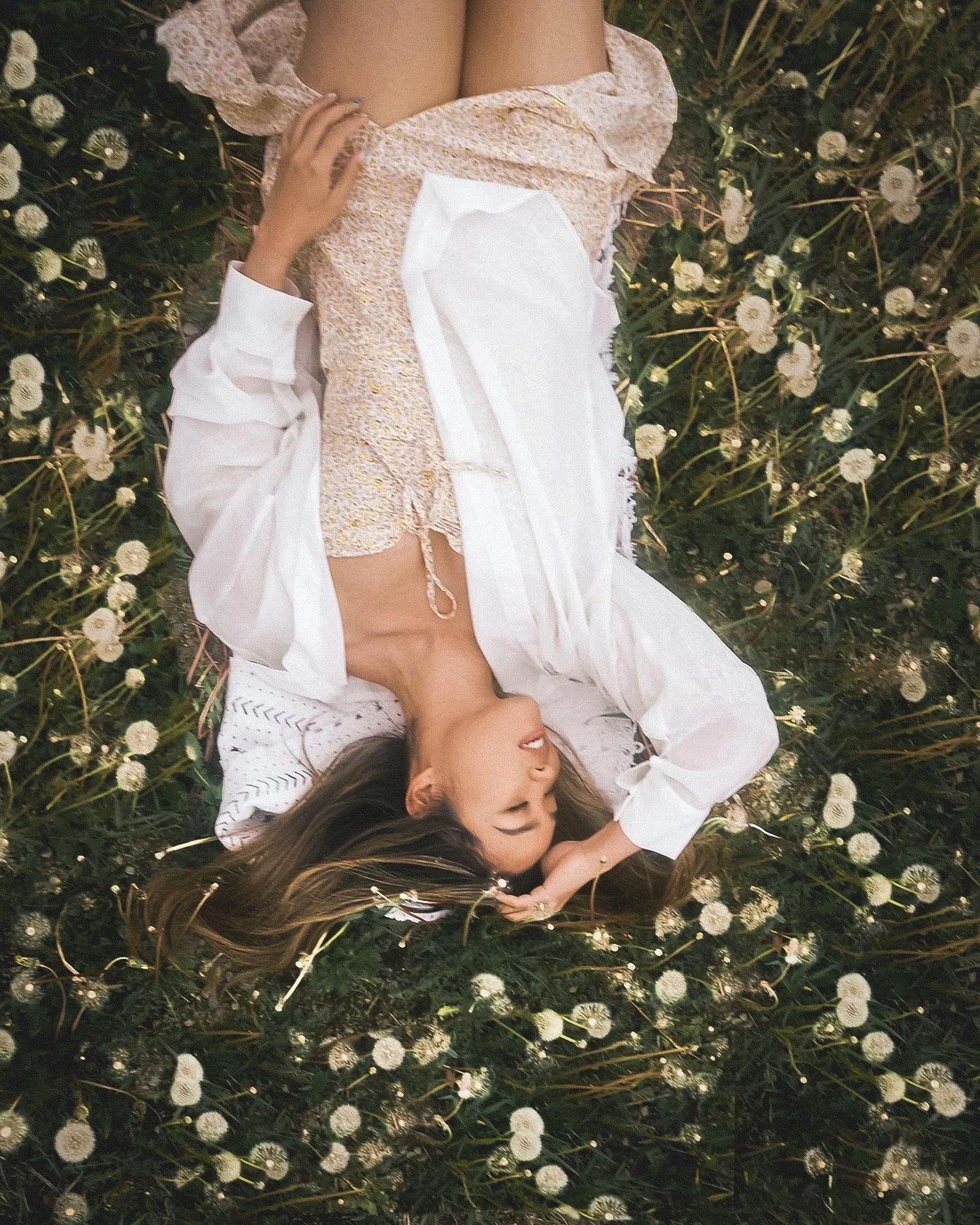 A woman lying on the ground in a field of white daisies, wearing a white shirt, a beige lace top, and beige shorts, with long brown hair spread around her head.