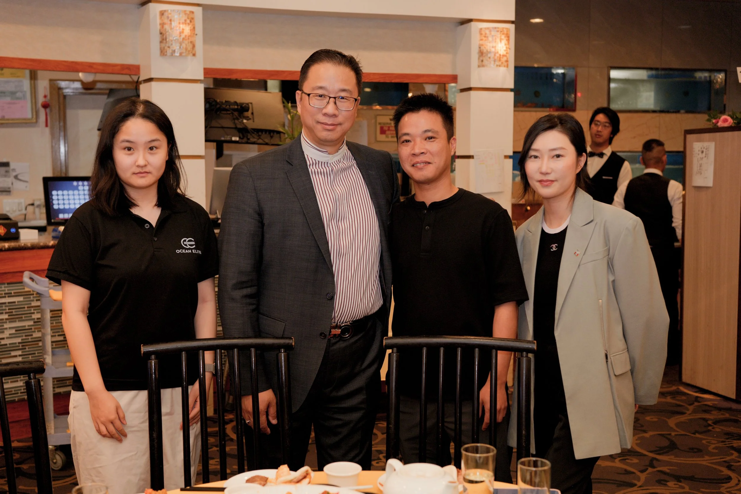 Four people standing together at a restaurant, posing for a photo. Two women on either side and two men in the middle, dressed in business casual attire. The background shows restaurant staff and decor.