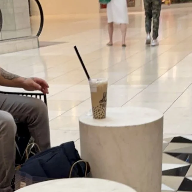 A plastic cup of bubble tea with a black straw on a white round table in a shopping mall, with people walking in the background.