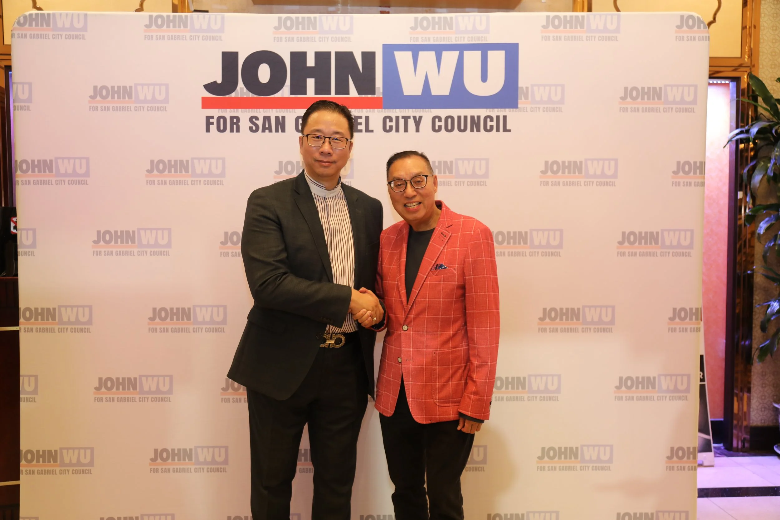 Two men shaking hands and smiling in front of a step and repeat banner with the text "John Wu for San Gabriel City Council". One man is wearing glasses, a dark suit, and a striped shirt. The other is wearing glasses and a pink checkered blazer.