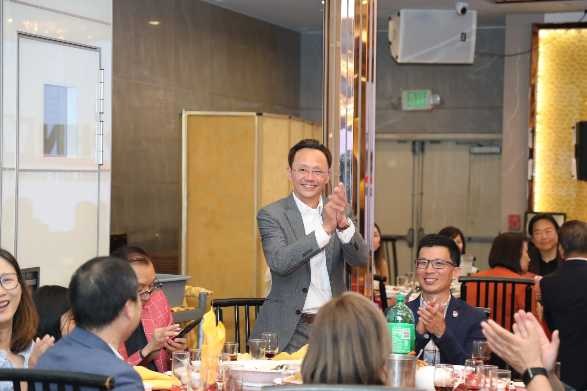 A man in a gray suit and glasses is standing and smiling at a gathering, surrounded by seated people, at a formal event or conference.