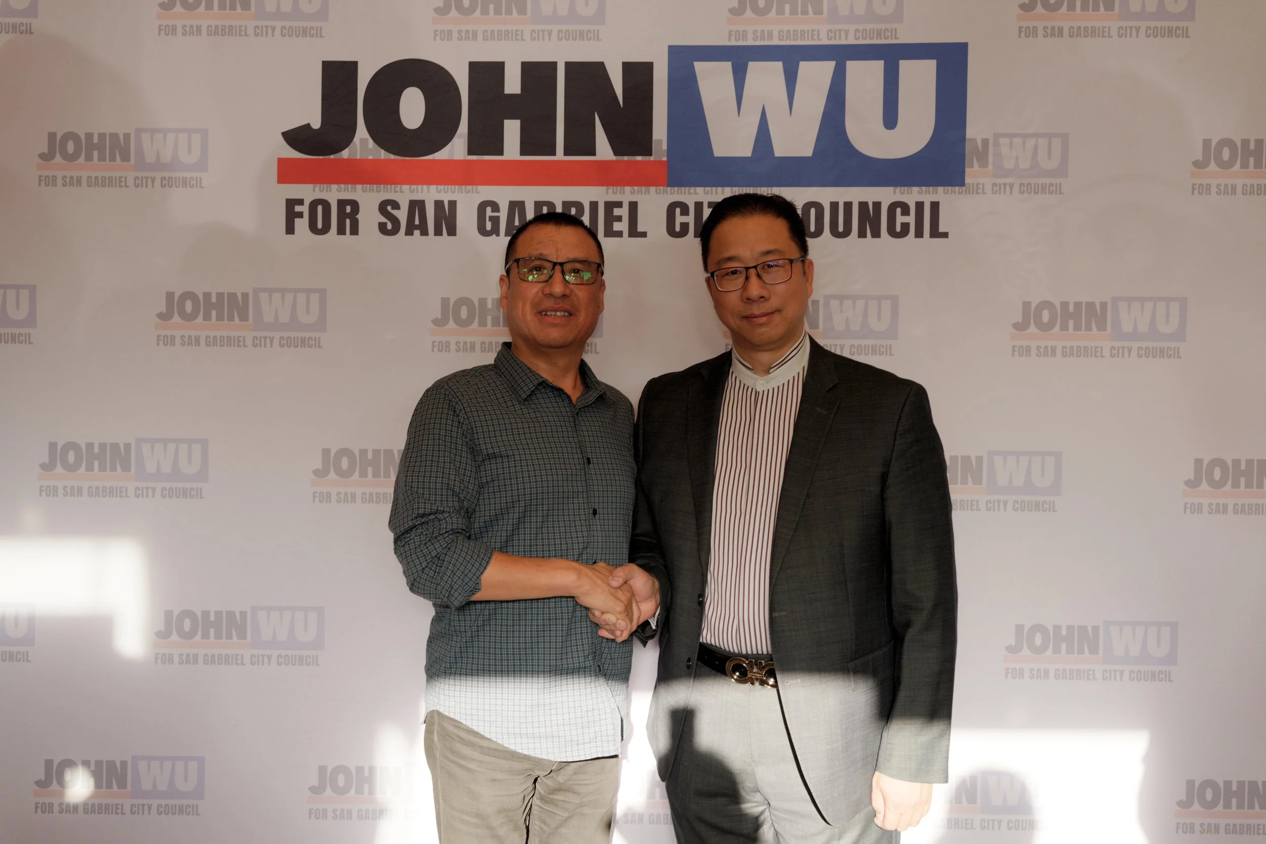 Two men in business attire shaking hands in front of a campaign backdrop with the text 'JOHN WU for San Gabriel City Council.'