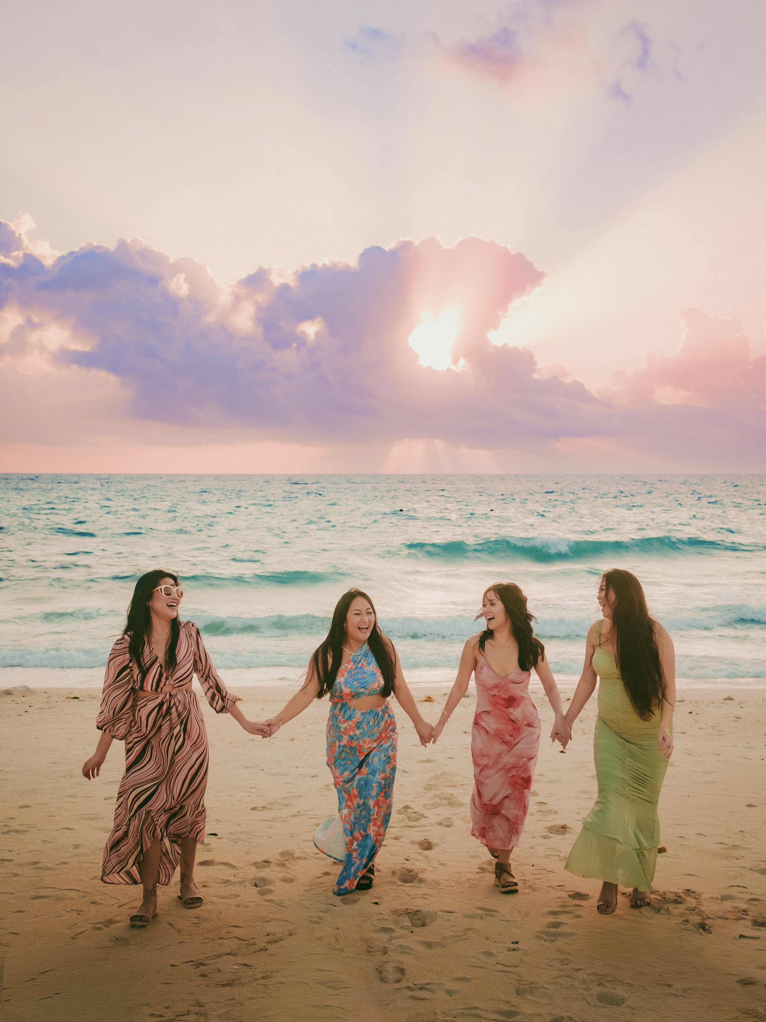 Four women holding hands and walking on the beach at sunset, smiling and laughing, wearing colorful dresses.