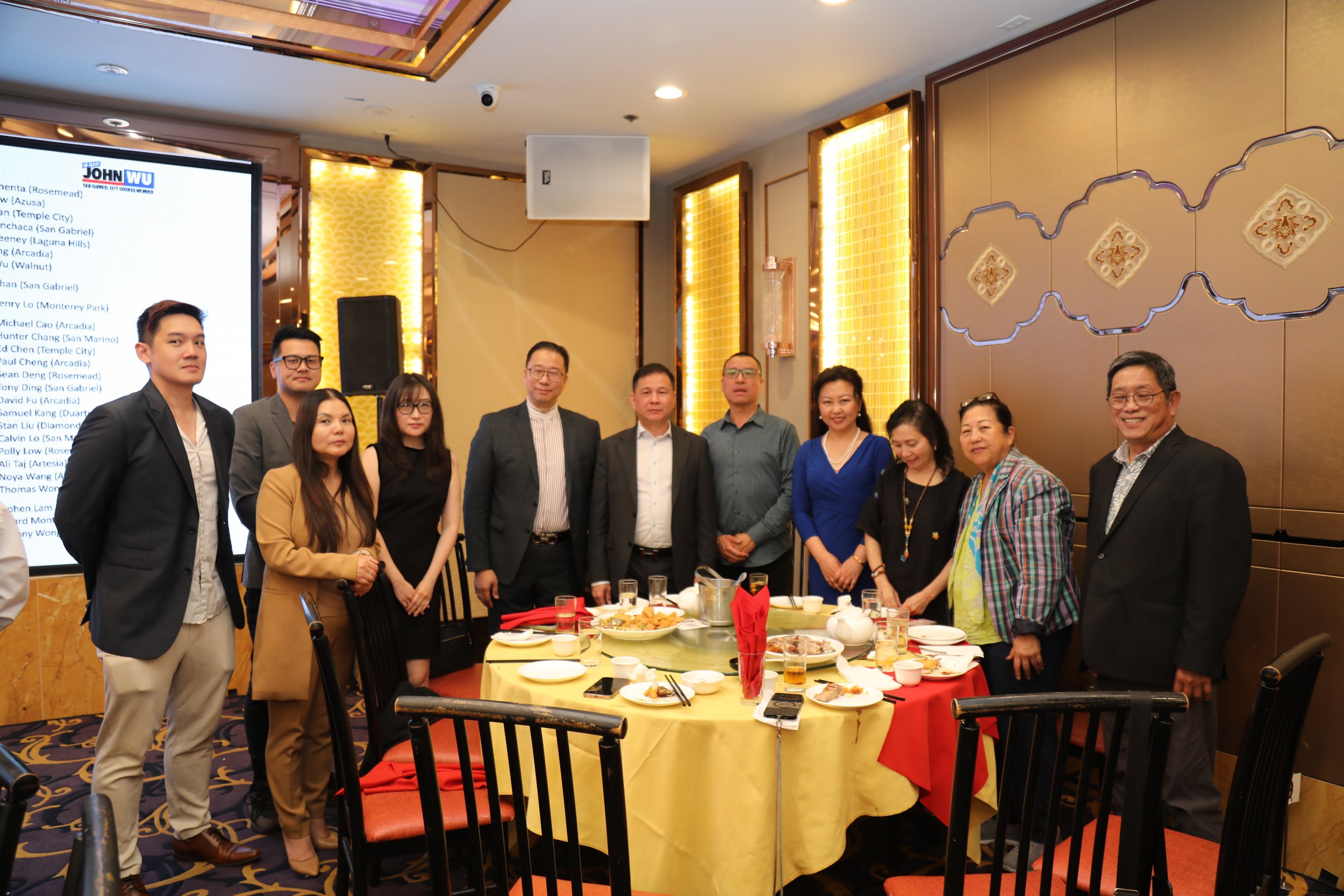 A group of eleven people standing around a dining table at a banquet or celebration, with food and drinks on the table, in an elegant room with decorative wall panels and a large screen displaying a list of names in the background.