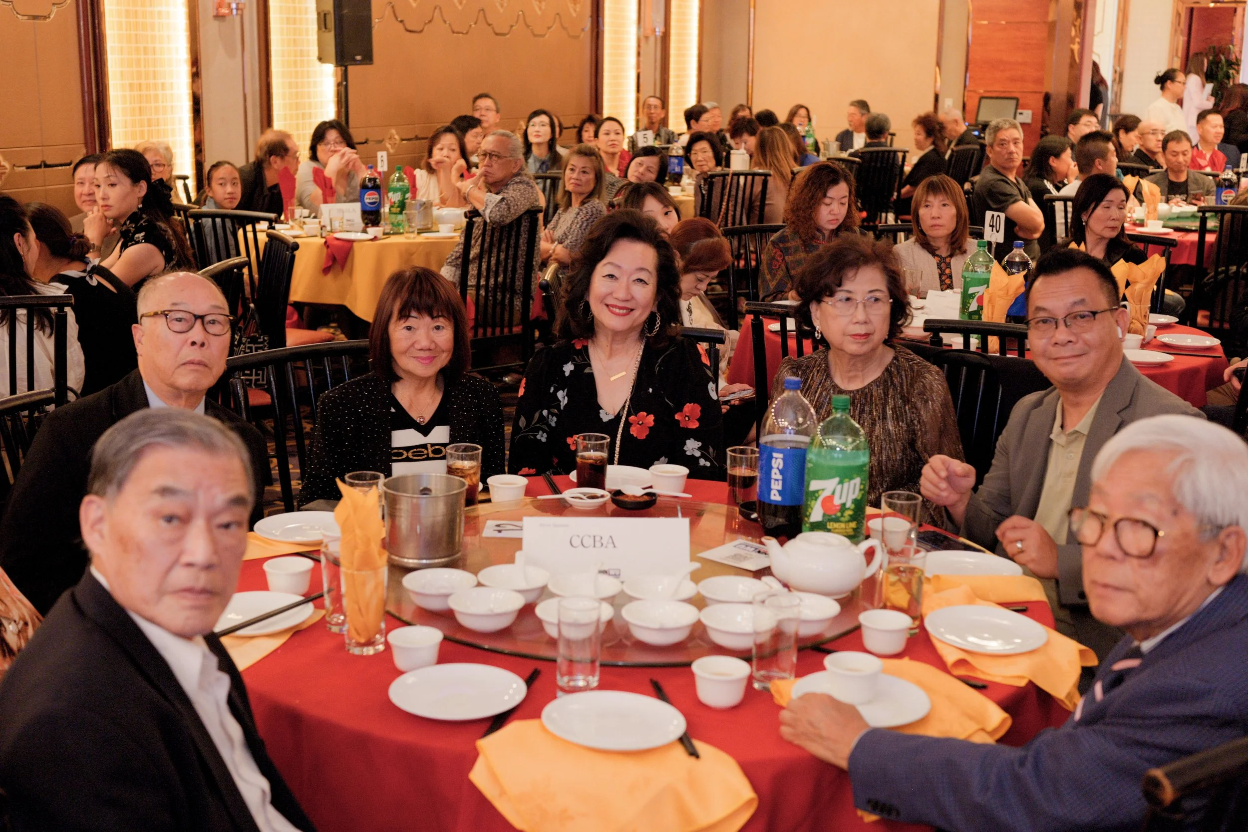 A large gathering of diverse people seated at round tables during a formal event or banquet, with some smiling and looking towards the camera. The tables are set with various dishes, bottles of soda, teapots, and orange napkins. The room features war