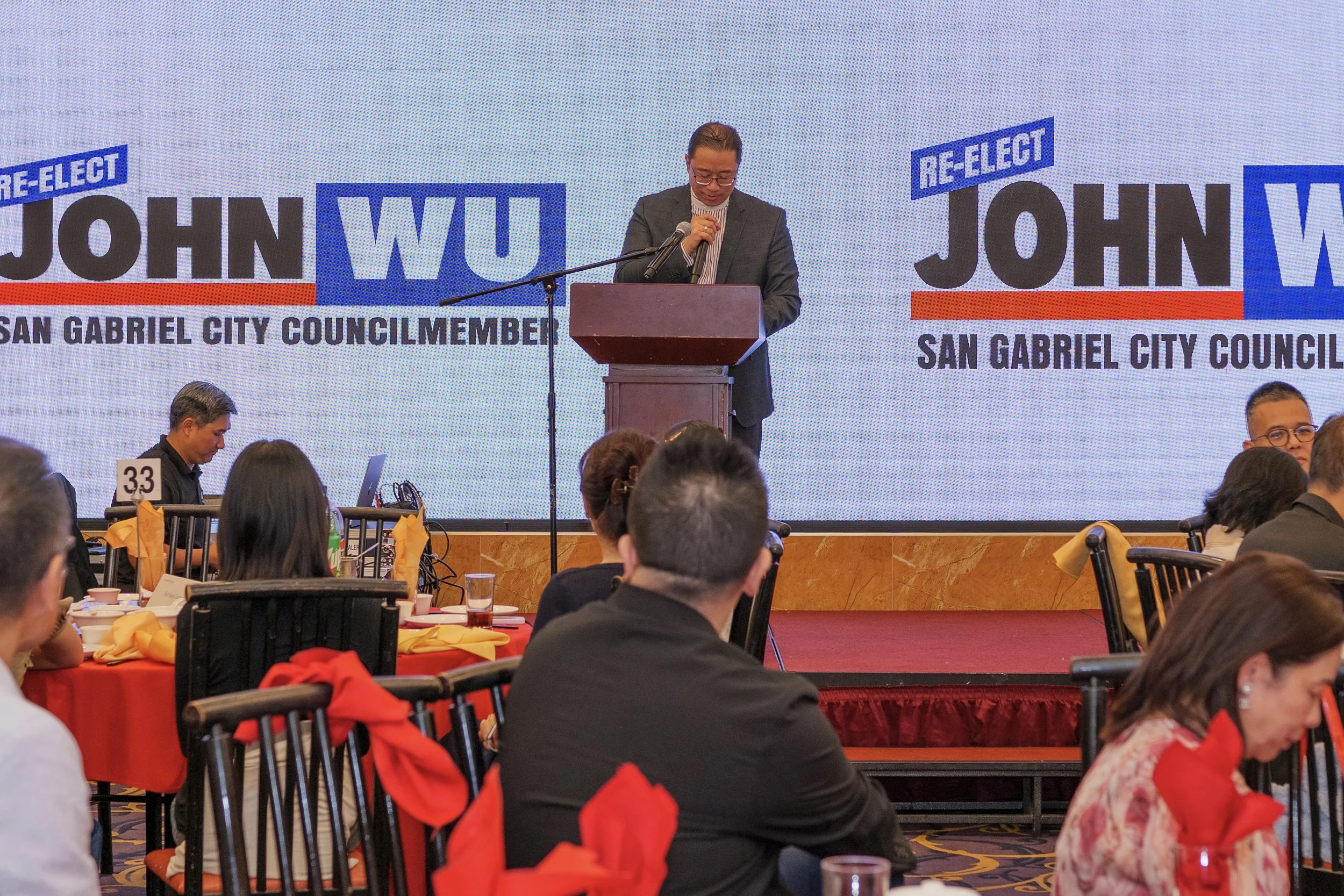 A man in a suit and glasses stands behind a podium, holding a piece of paper, with a large screen behind him displaying the words "RE-ELECT JOHN WU" and "SAN GABRIEL CITY COUNCILMEMBER". Several round tables with orange and red tablecloths, glasses, 