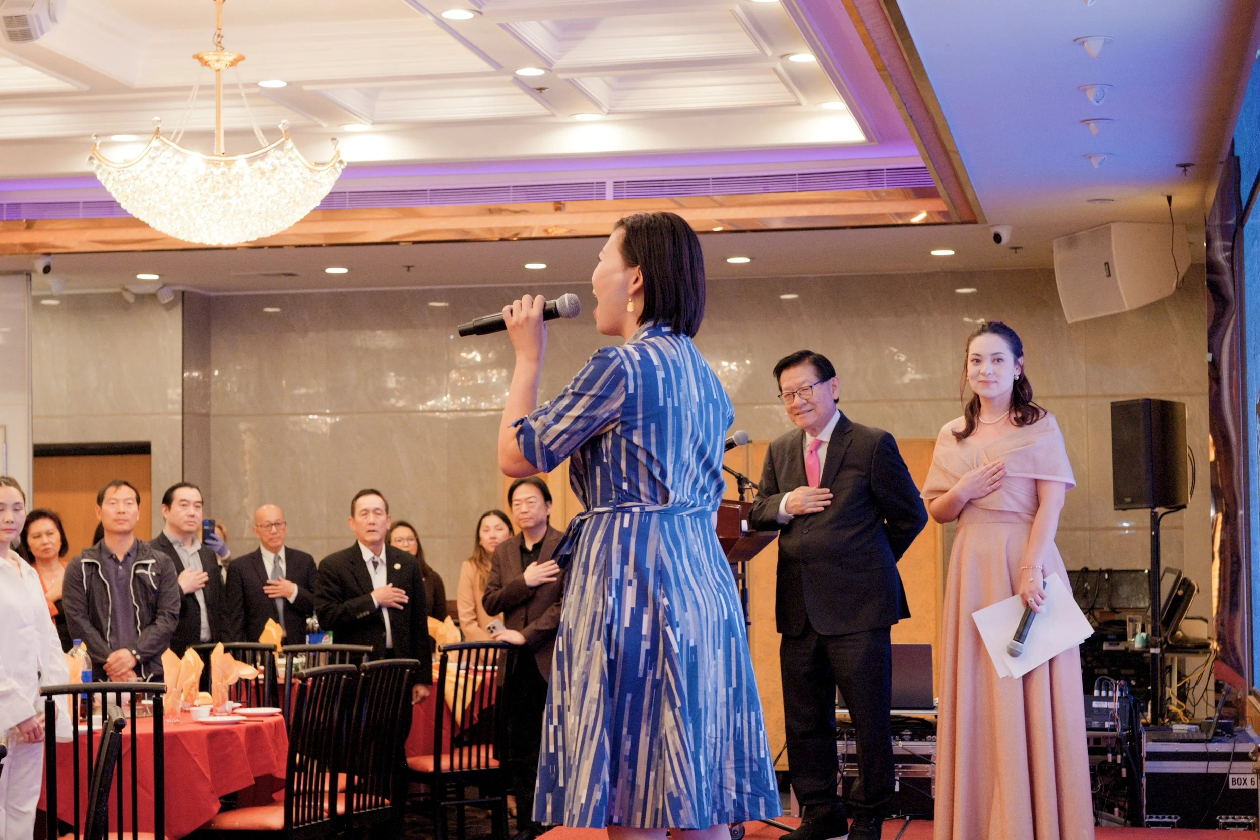 Woman in a blue patterned dress speaking into a microphone during a formal event, with audience members and a man and woman standing on stage in the background.