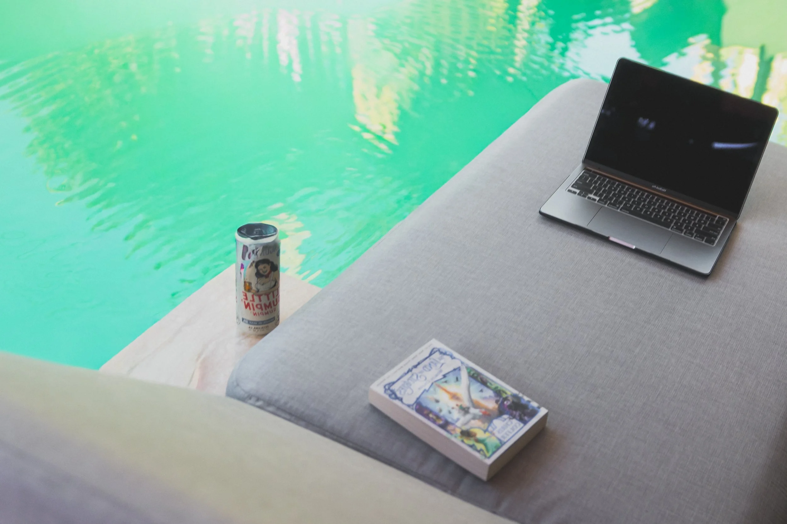 Laptop, paperback book and a drink can on the gray Solaris chaise lounge beside a bright green pool