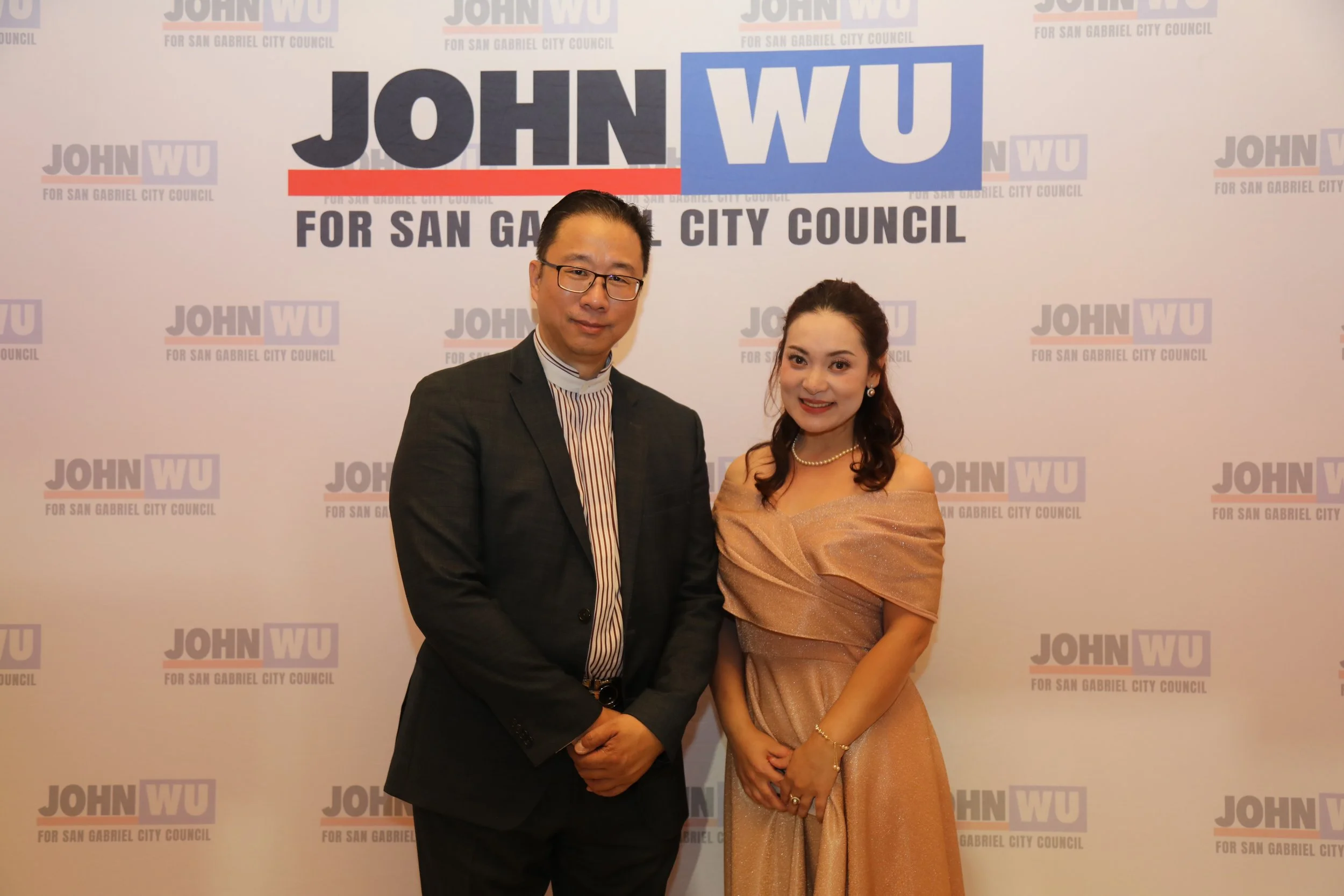 A man in a dark suit with glasses and a woman in a peach-colored dress standing together in front of a campaign backdrop for John Wu for San Gabriel City Council.
