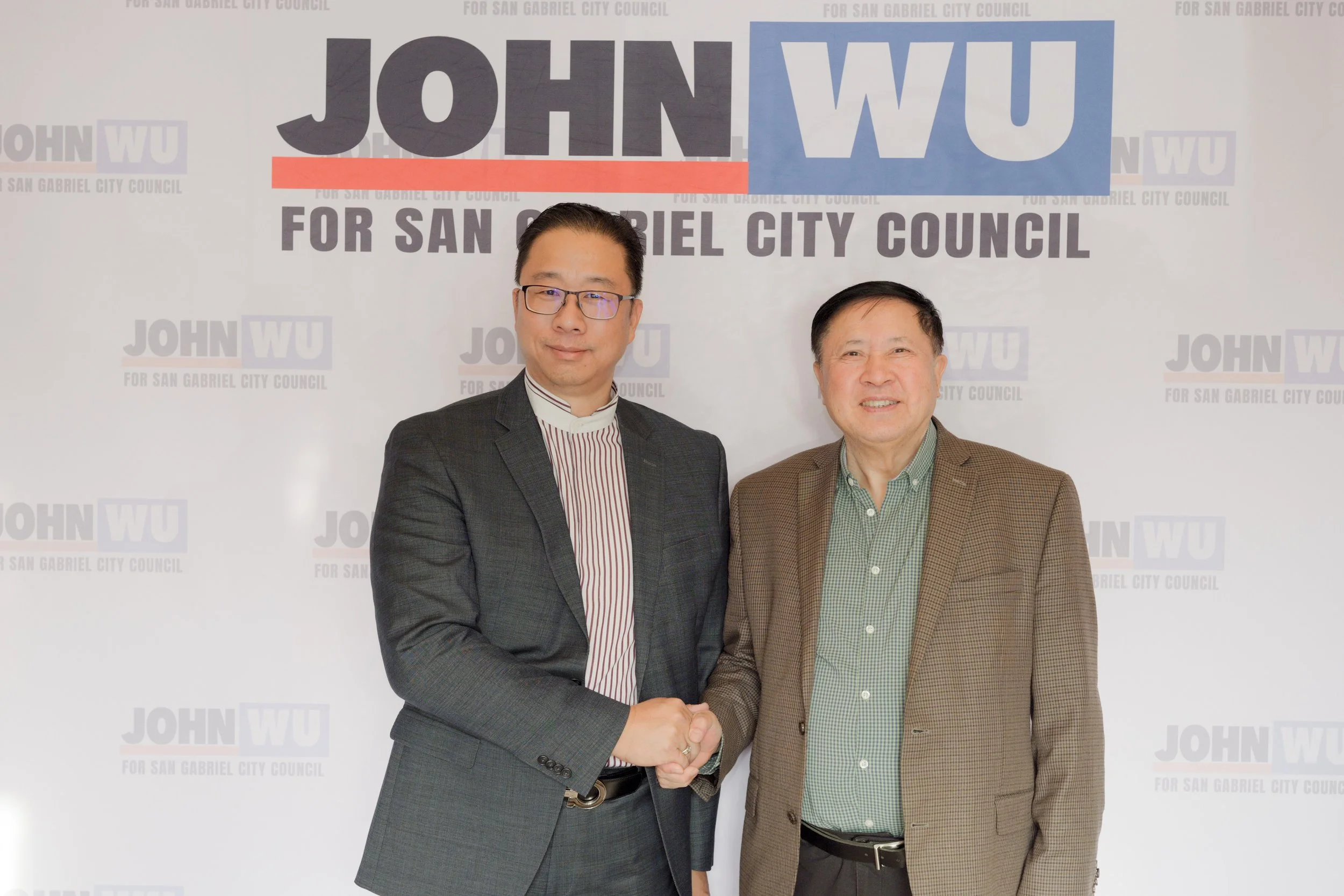 Two men in suits shaking hands in front of a backdrop supporting a campaign for San Gabriel City Council, featuring the name 'John Wu' in large block letters and the text 'For San Gabriel City Council'.