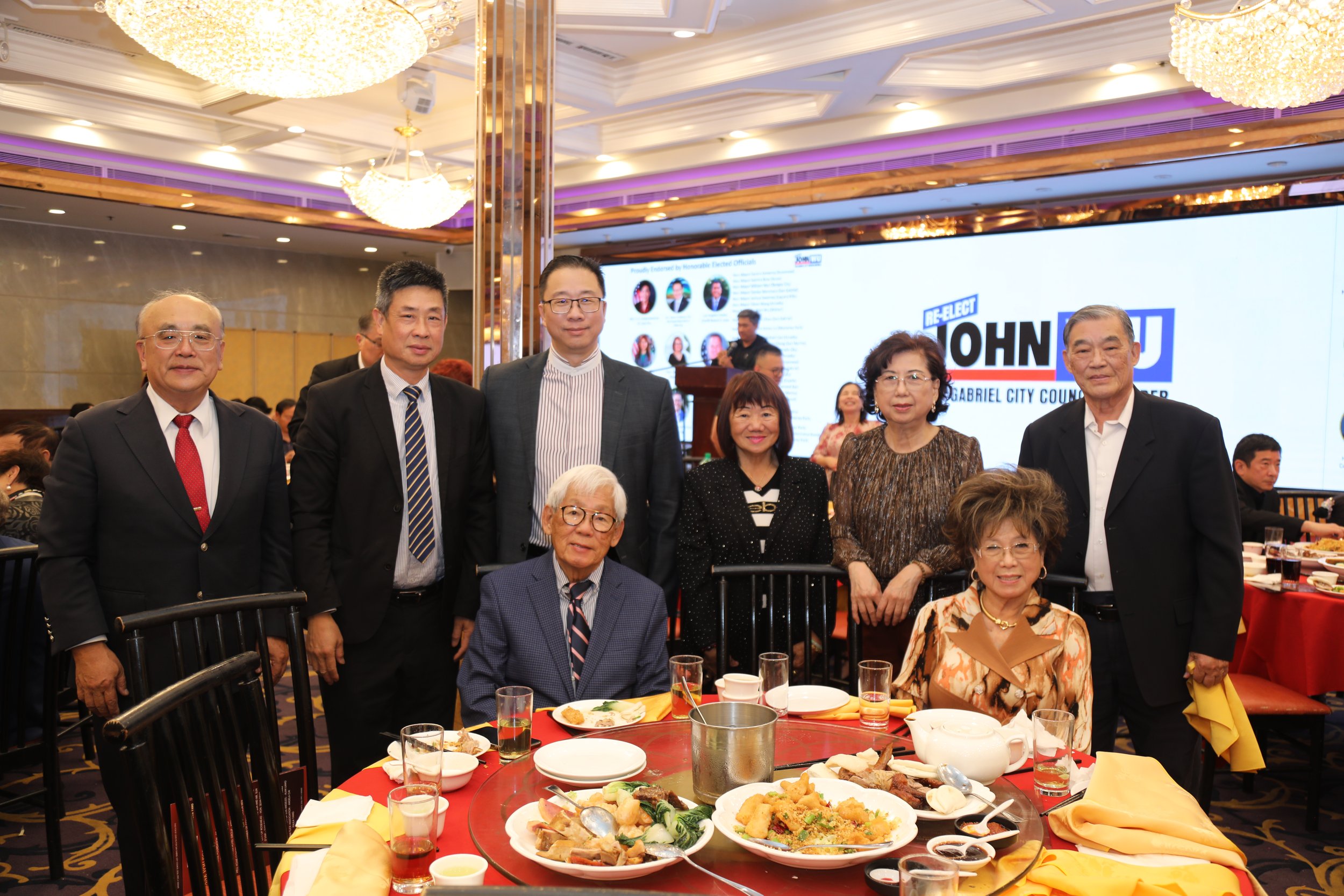 Group of Asian and Caucasian people at a banquet table with food, in a banquet hall with chandeliers and a large screen displaying political campaign information.