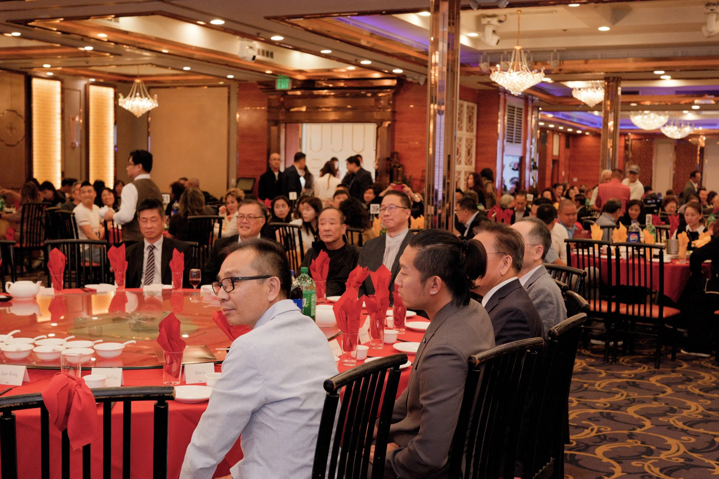 A formal banquet scene with many people seated at round tables decorated with red napkins and tablecloths, in a richly decorated hall with chandeliers and wooden accents.
