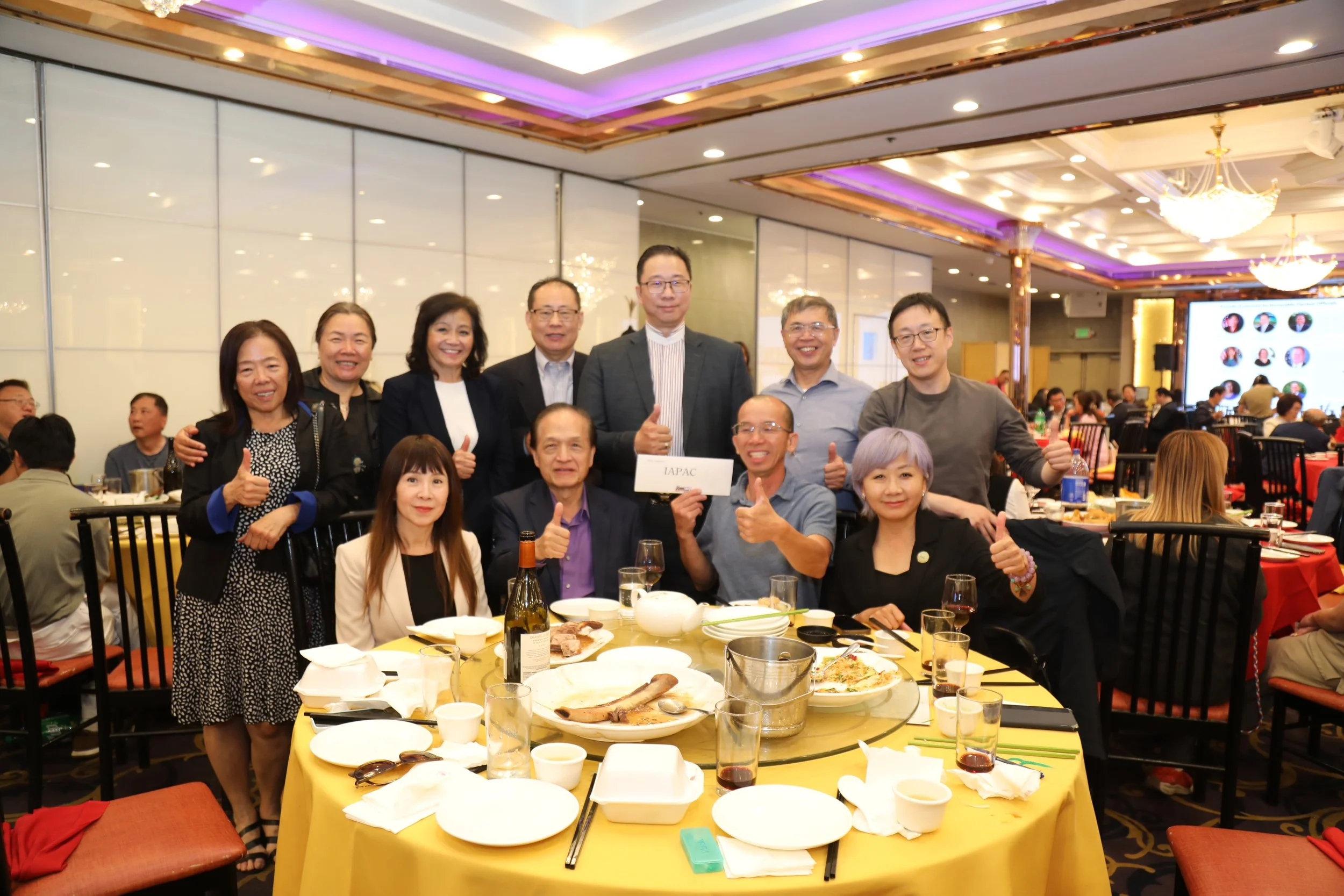 Group of people posing at a banquet table during an event in a decorated banquet hall, with food and drinks on the table, some people giving thumbs up, and a large screen in the background showing profile pictures.