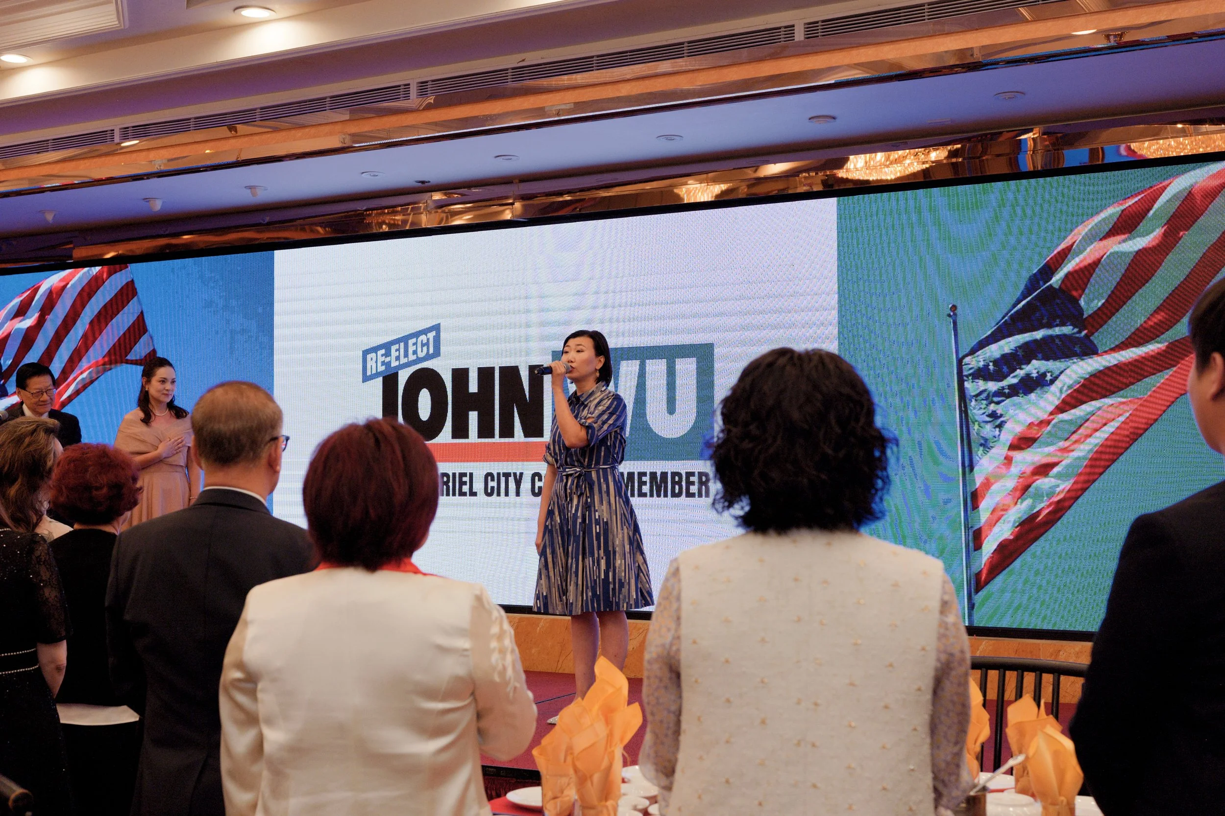 A woman speaking into a microphone at a political event with a large screen behind her displaying the words 'Re-elect Johyu'. The audience is seated, and the setting appears to be a formal indoor gathering with American flags on display.