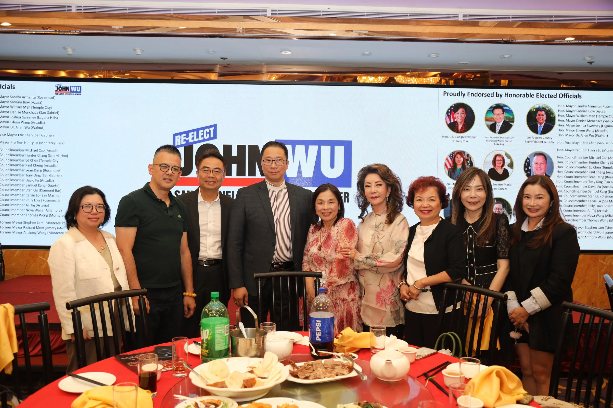 A group of people standing together at a banquet table during a formal event, with a large screen behind them displaying political endorsements and the word 'RE-ELECT' in blue. The table is set with various dishes, snacks, and drinks.