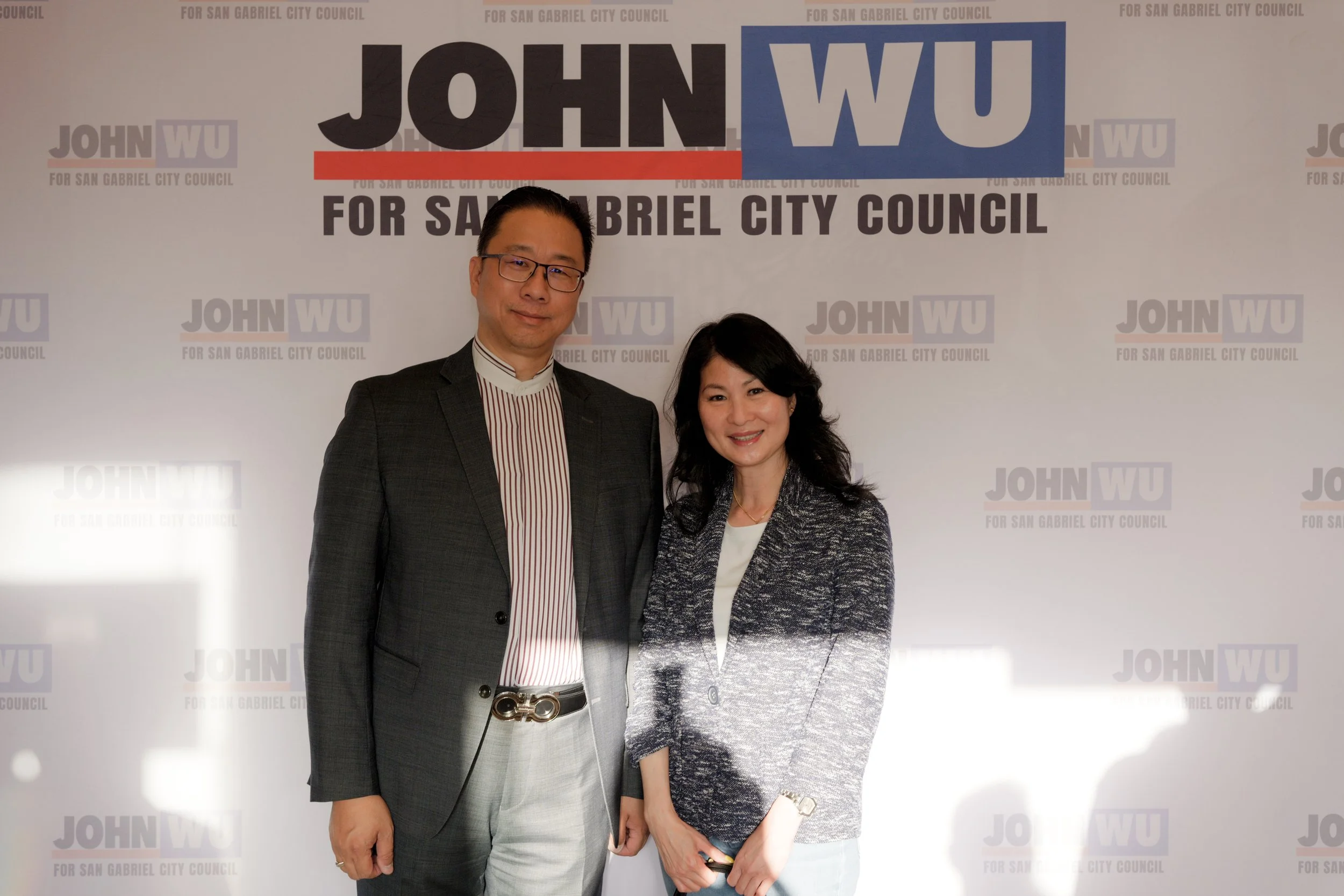 Two people standing in front of a backdrop with campaign signs that say 'JOHN WU for San Gabriel City Council'. The man on the left is wearing glasses, a dark plaid blazer, a white shirt with red stripes, and a belt with a distinctive buckle. The wom