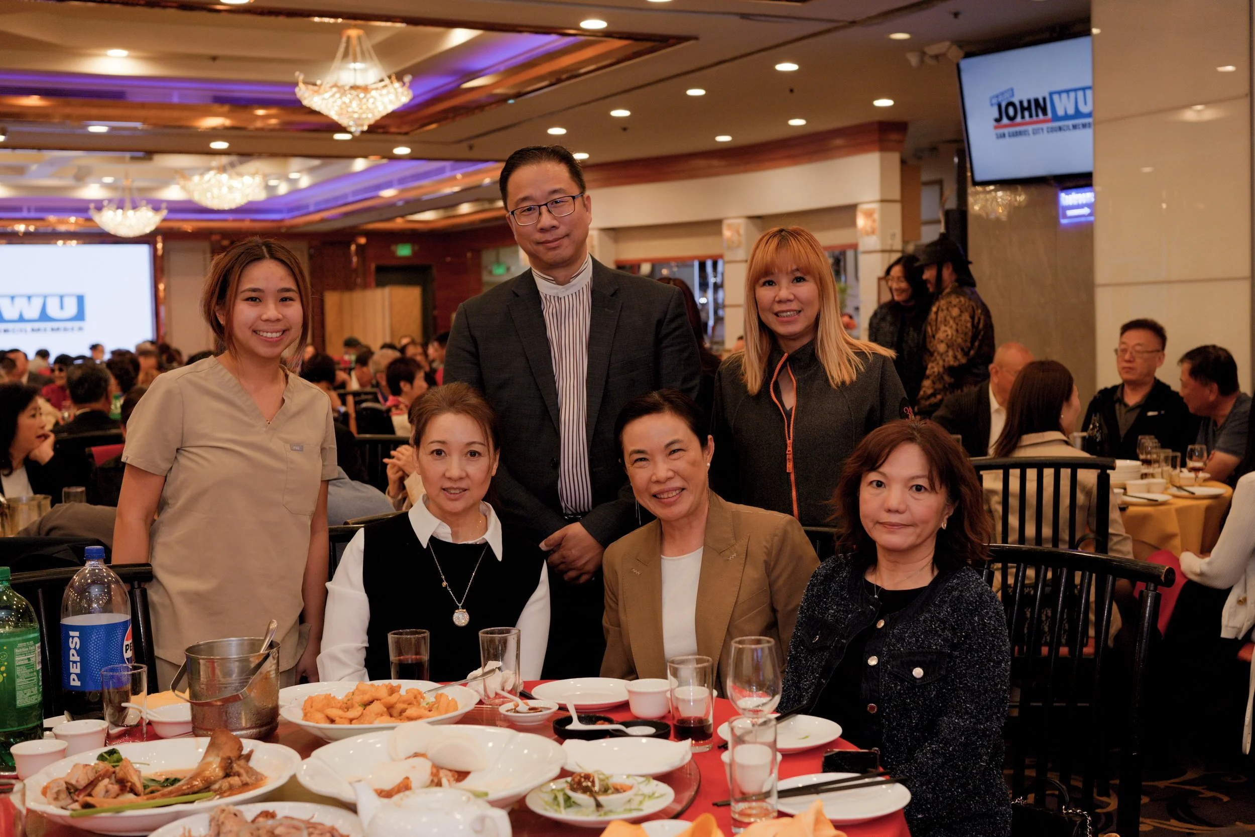 A group of five people posing for a photo at a banquet or dinner event, with tables of food in front of them and a large screen in the background displaying the logo for a political campaign.