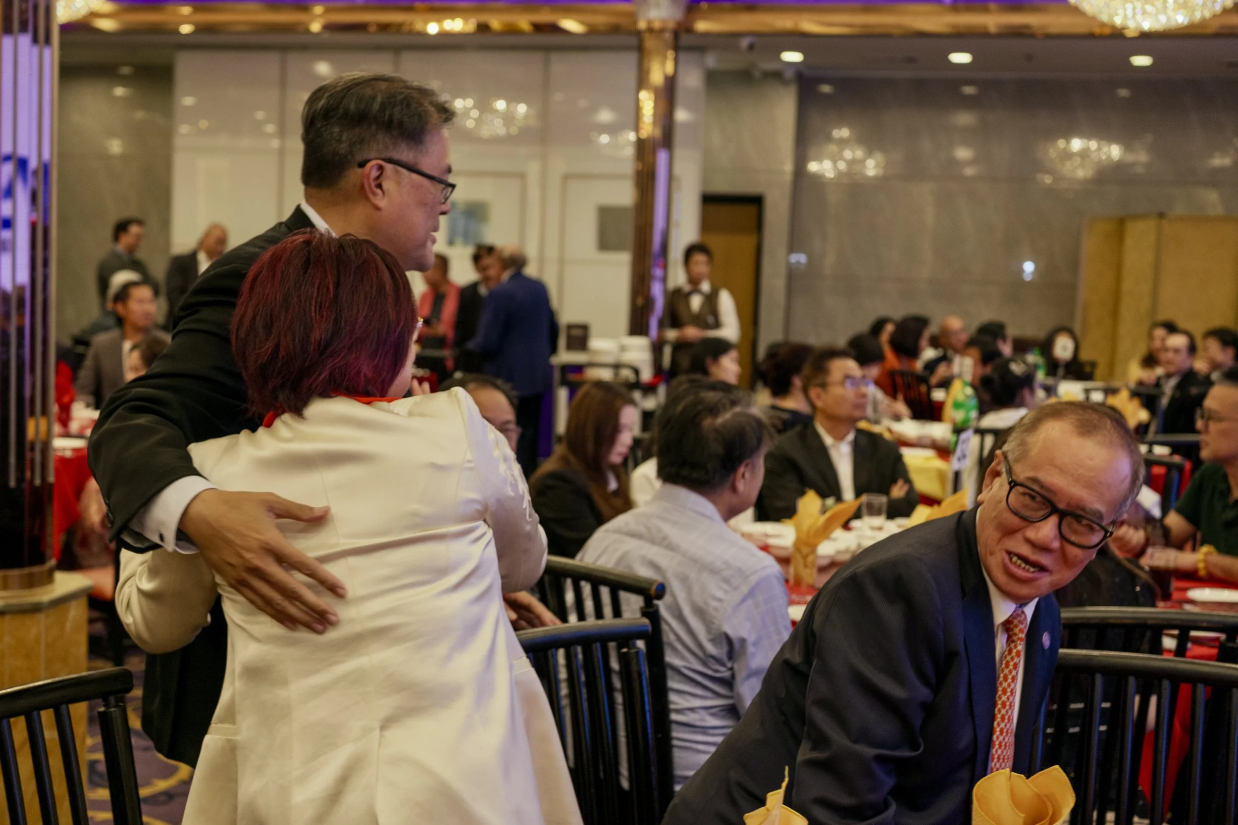 People seated at a banquet table in a conference or event setting, with a woman standing and embracing a man in a suit, blurred background of other attendees and wait staff.