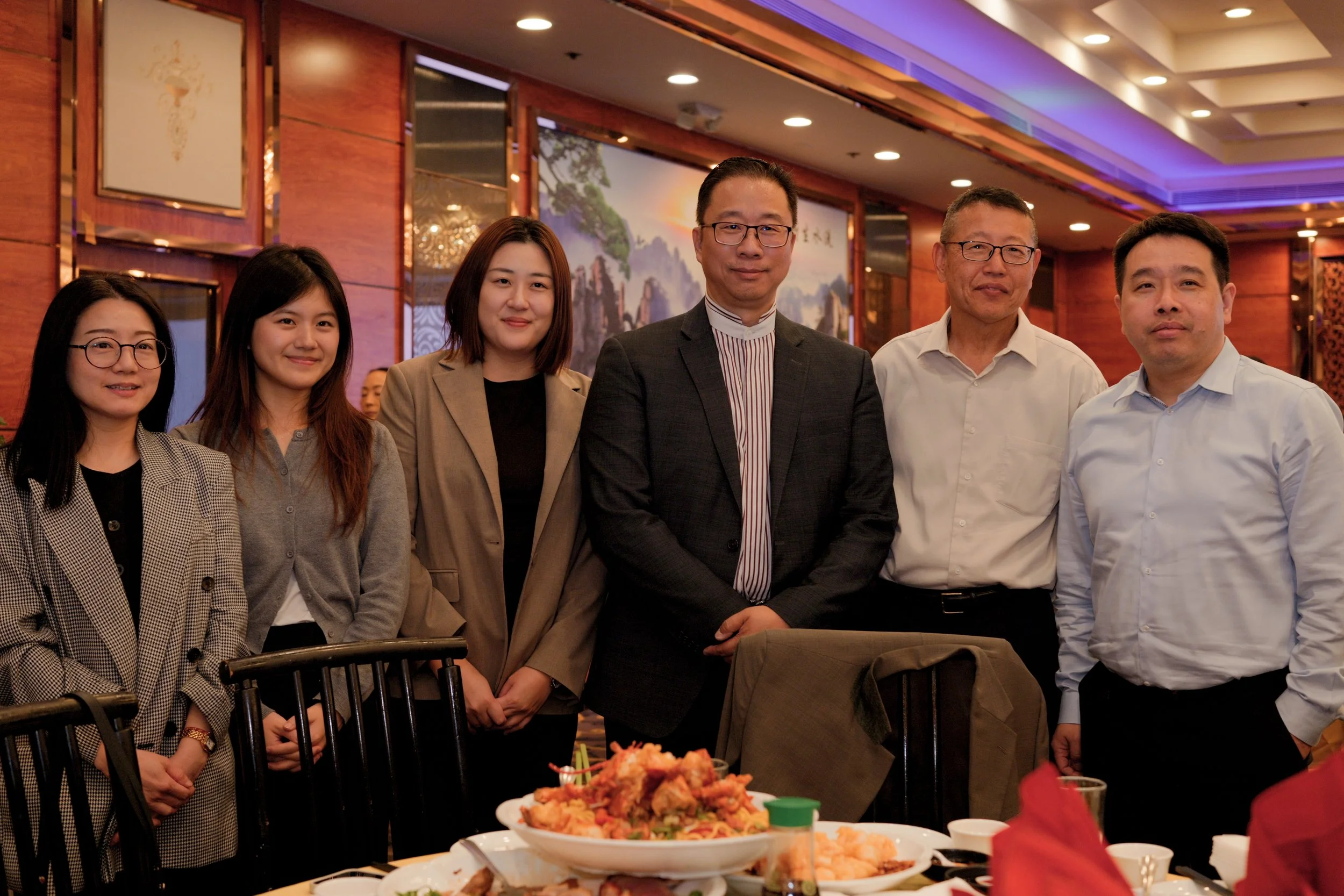 Group of six diverse people standing together at a formal gathering in a decorated restaurant or banquet hall.
