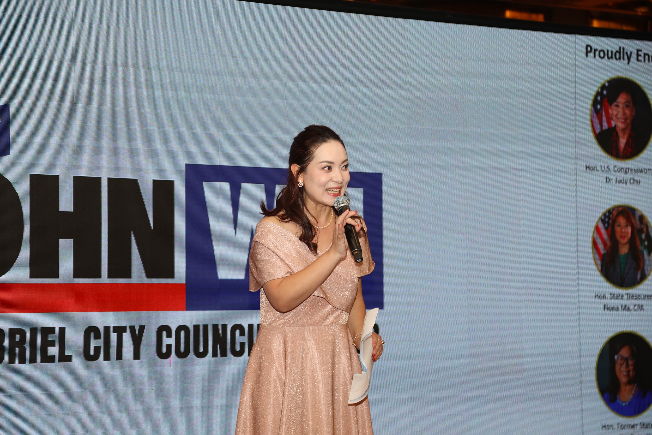 A woman in a peach-colored dress speaking into a microphone at a conference, with a large screen displaying her photo and text in the background.