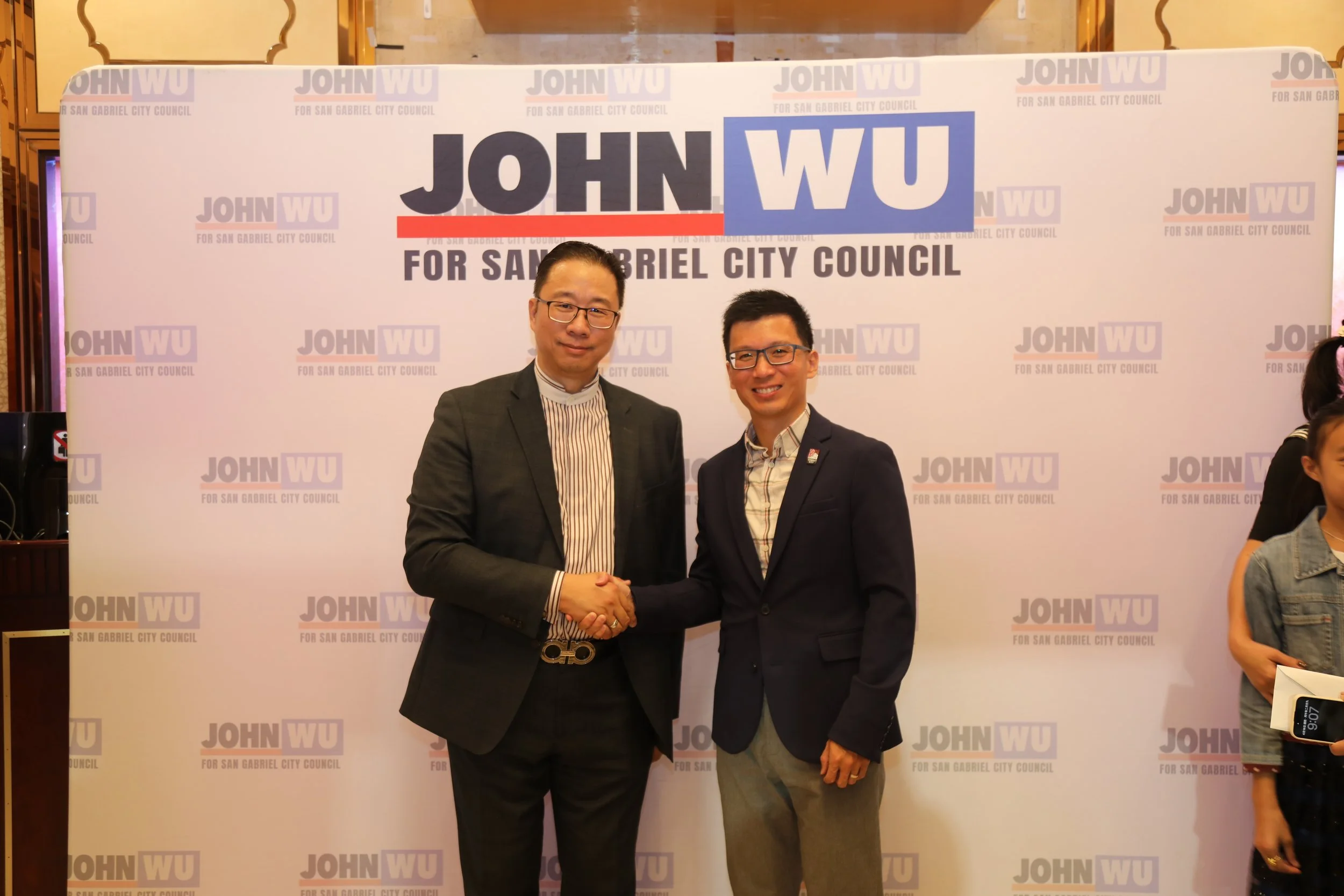 Two men shaking hands in front of a campaign banner that says "JOHN WU for San Gabriel City Council." One man is wearing glasses, a gray plaid suit, and a striped shirt. The other man is wearing glasses, a dark blazer, and a light shirt.