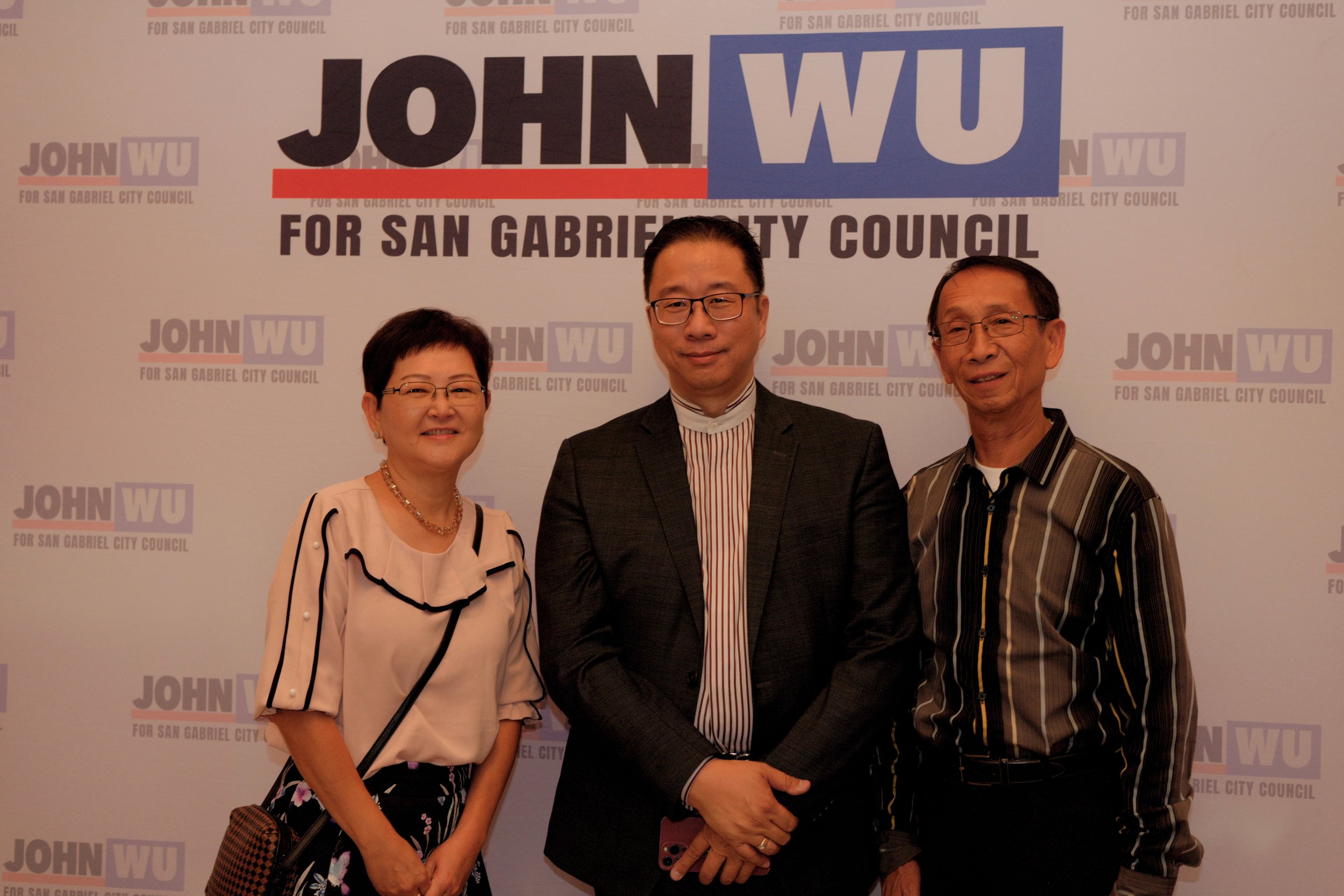 Three people standing in front of a backdrop with campaign logos for John Wu for San Gabriel City Council. The woman on the left is wearing glasses, a light-colored blouse with black trim, and a pearl necklace. The man in the middle, wearing glasses 