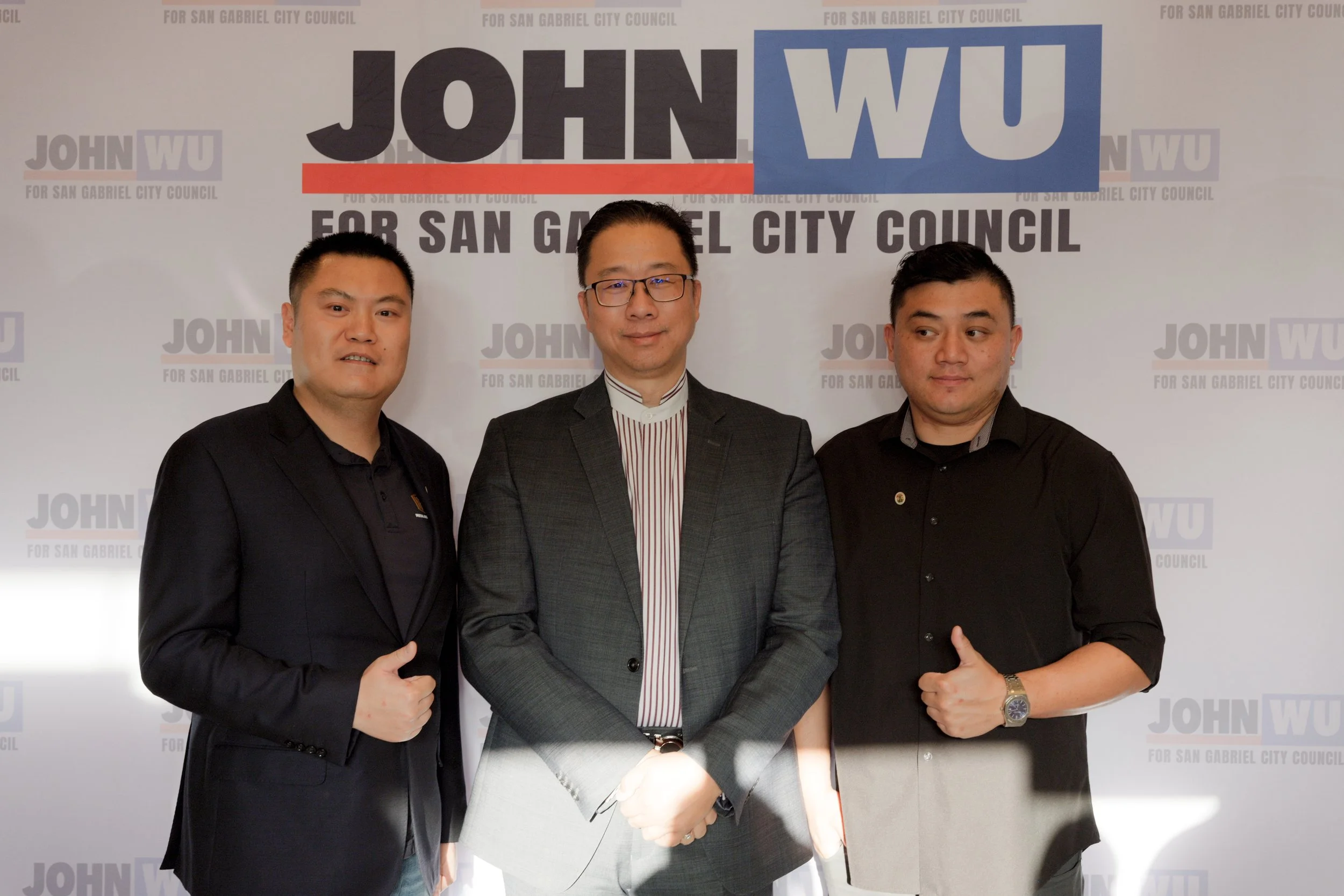 Three men standing together in front of a white backdrop with the text 'JOHN WU FOR SAN GABRIEL CITY COUNCIL' repeated in a pattern. The man in the middle is wearing glasses and a suit, while the men on either side are wearing dark shirts, giving a t