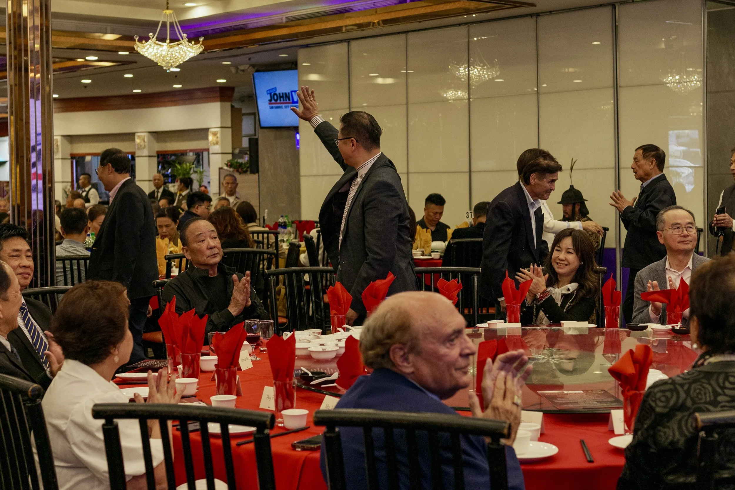 A banquet hall filled with people seated at round tables with red tablecloths, red folded napkins, and dinnerware, while some individuals stand and converse. A man in a suit and glasses is waving, and a woman with long hair and a man with dark hair a