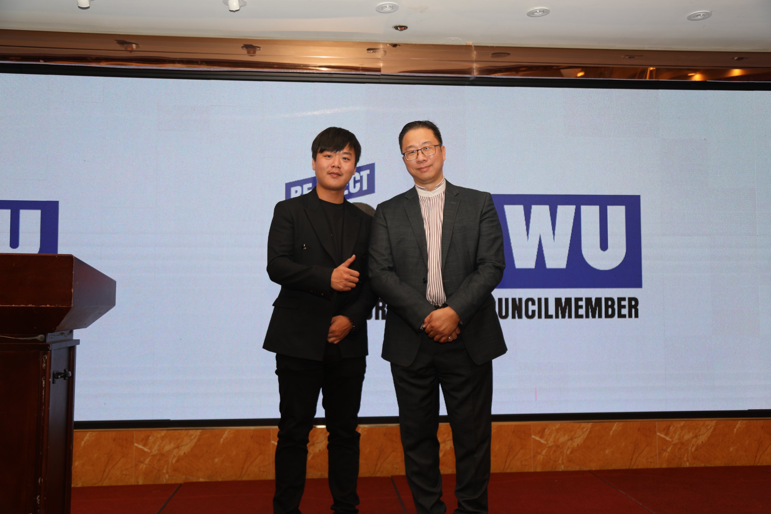Two men standing together on a stage in front of a screen with the text 'REACT WU' and 'COUNCIL MEMBER', dressed in business suits, looking at the camera.