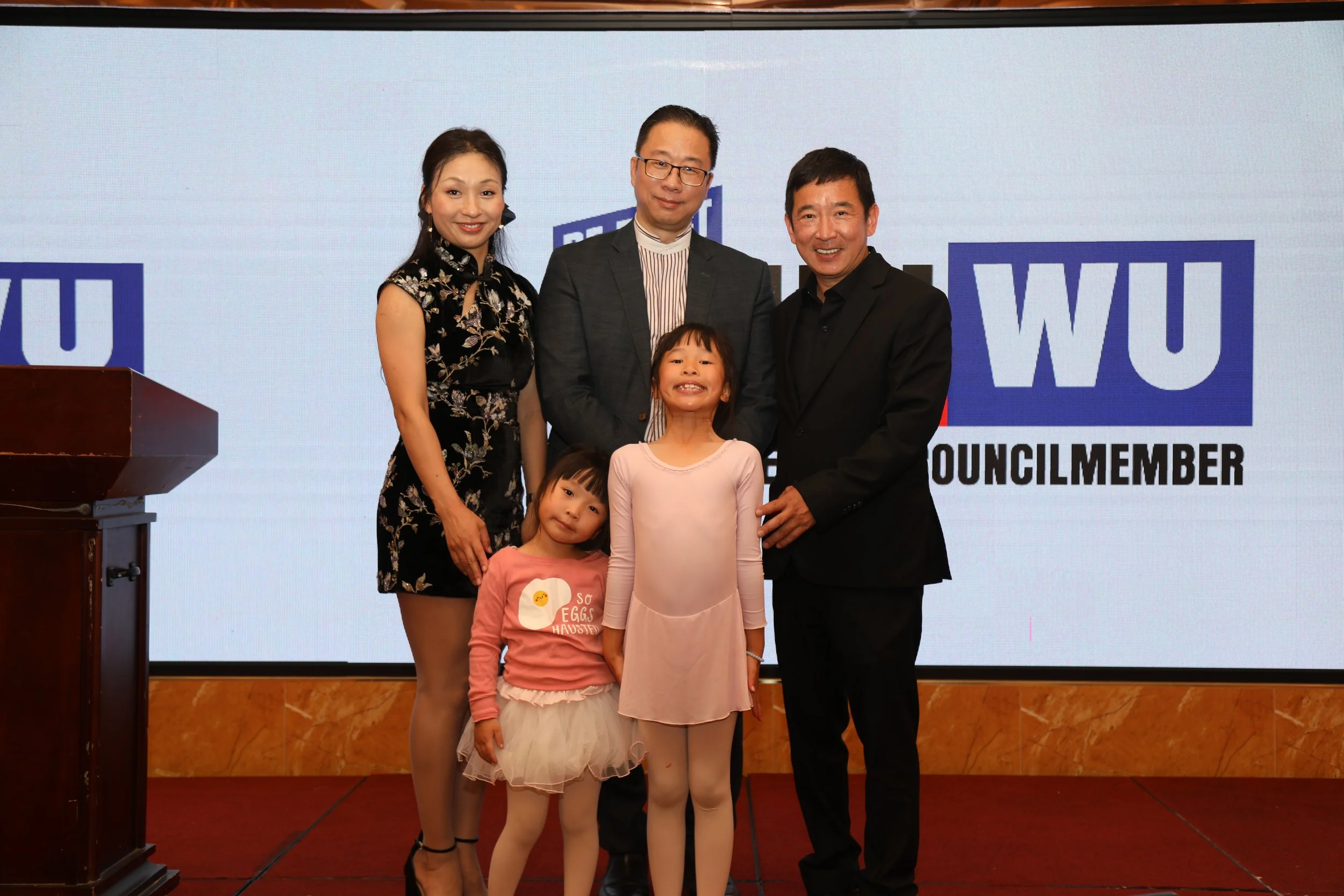 Group of three adults and three children posing for a photo in front of a large screen displaying the word 'Councillor.' The adults are standing behind the children, smiling, with two men in suits and a woman in a floral dress. The children are in fr