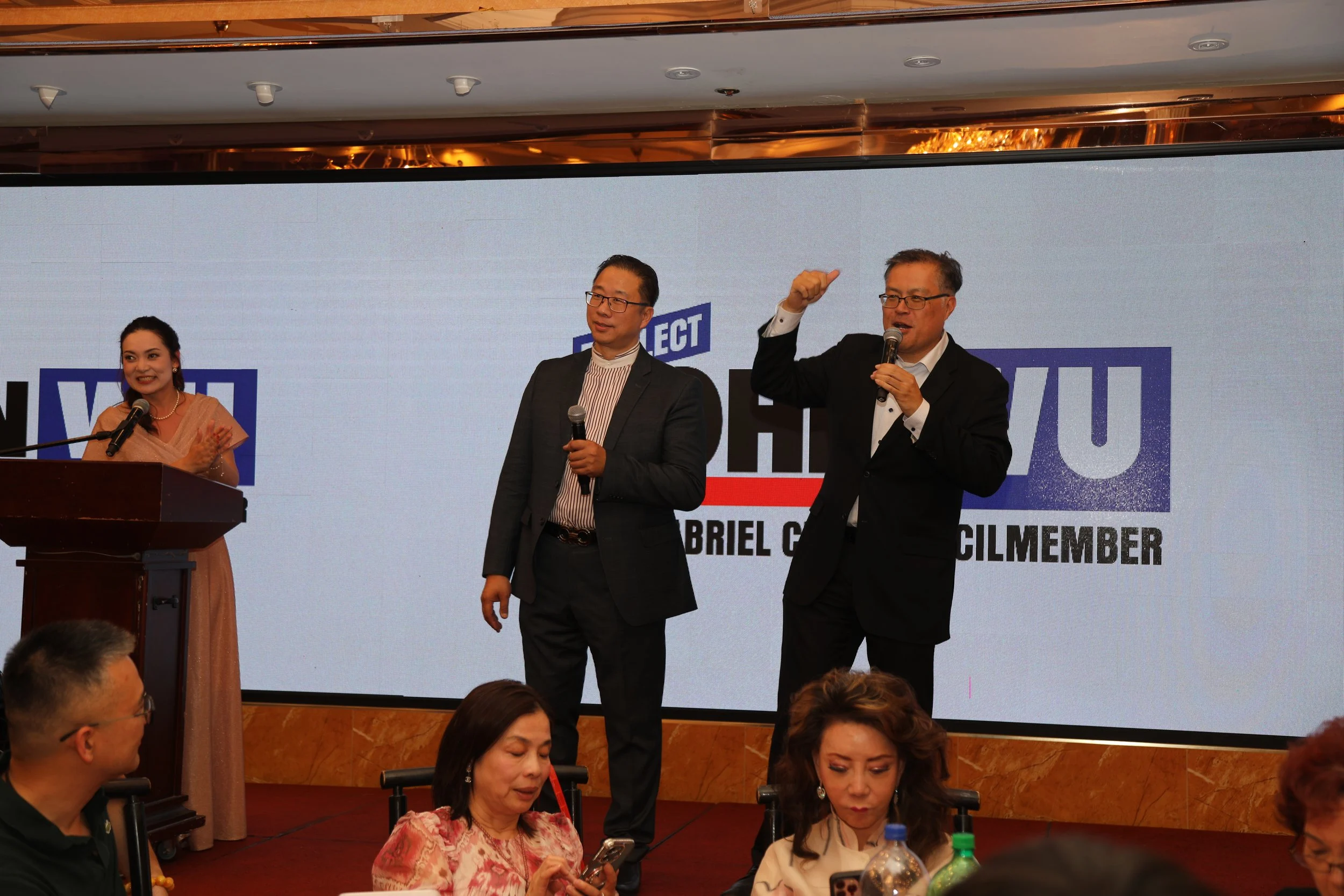 Three men and two women are at a formal event, with the men standing on stage in front of a large screen. The woman at the podium is speaking into a microphone, and the men are holding microphones, with one gesturing with his hand. The large screen b